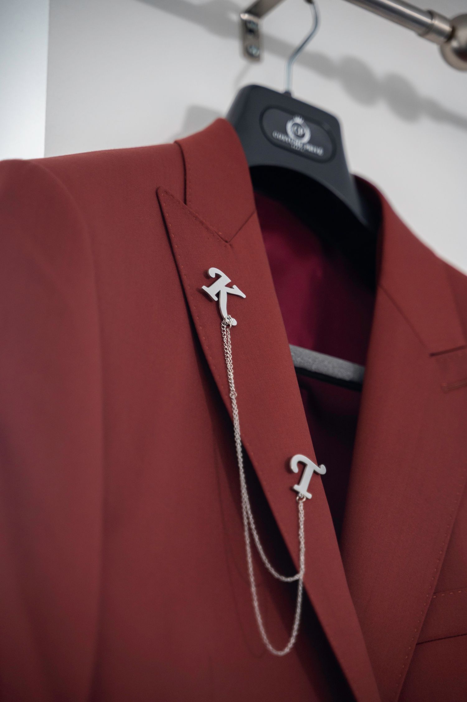 Burgundy suit jacket on hanger with silver K and T monogram chain lapel pins.