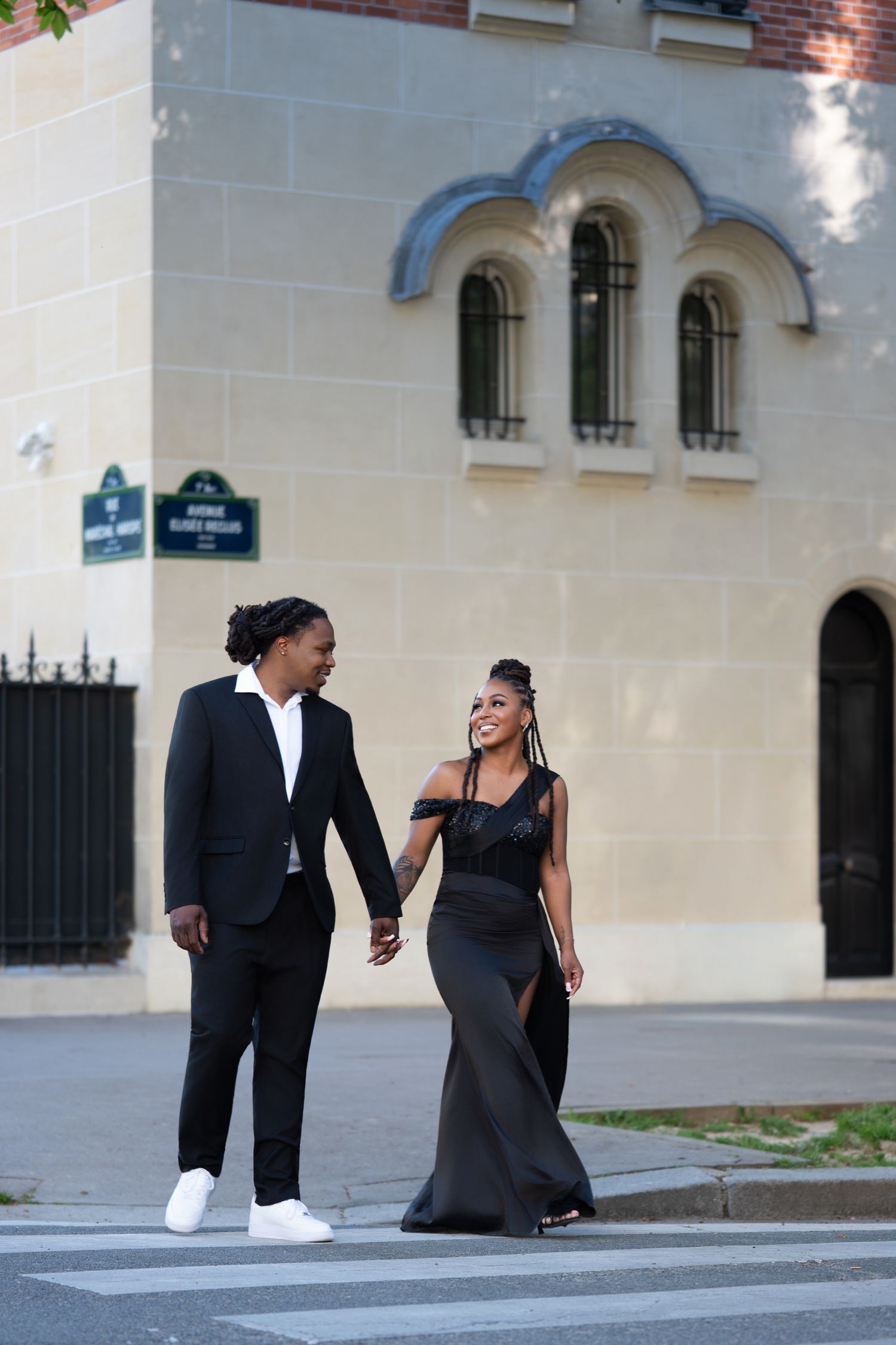 Elegant couple in black formal wear holding hands while crossing a street in Paris near a classic stone building.
