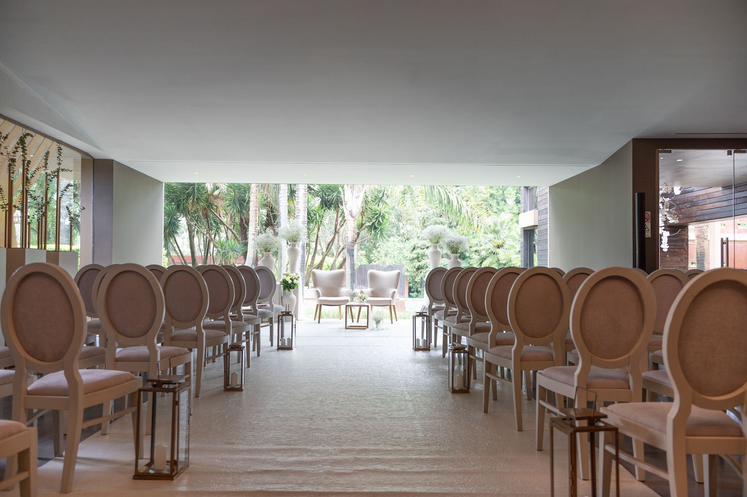 Elegant wedding ceremony venue with rows of beige round-back chairs leading to a garden backdrop.