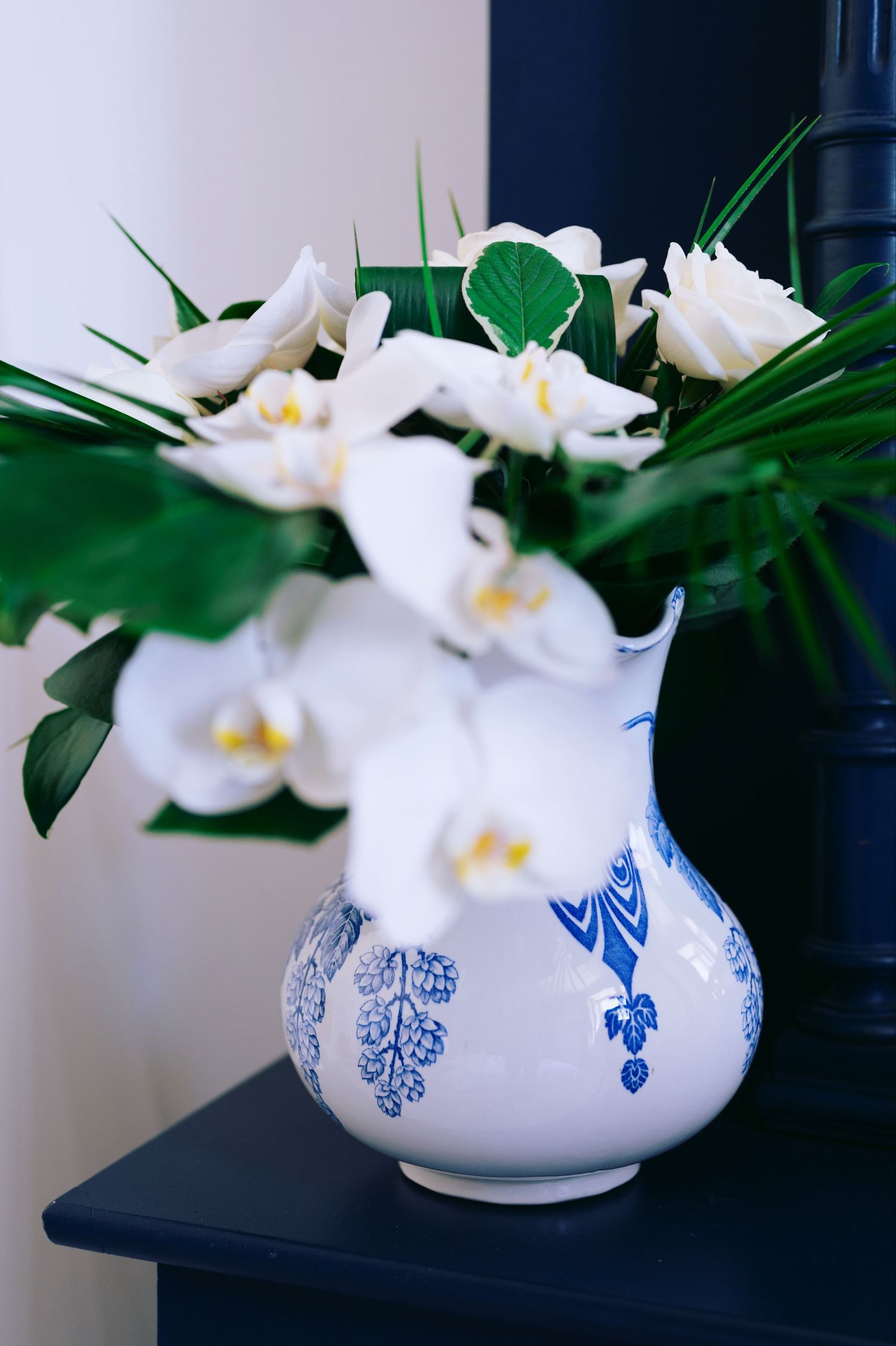 White and blue porcelain vase with white orchids and tropical green leaves on a dark surface.