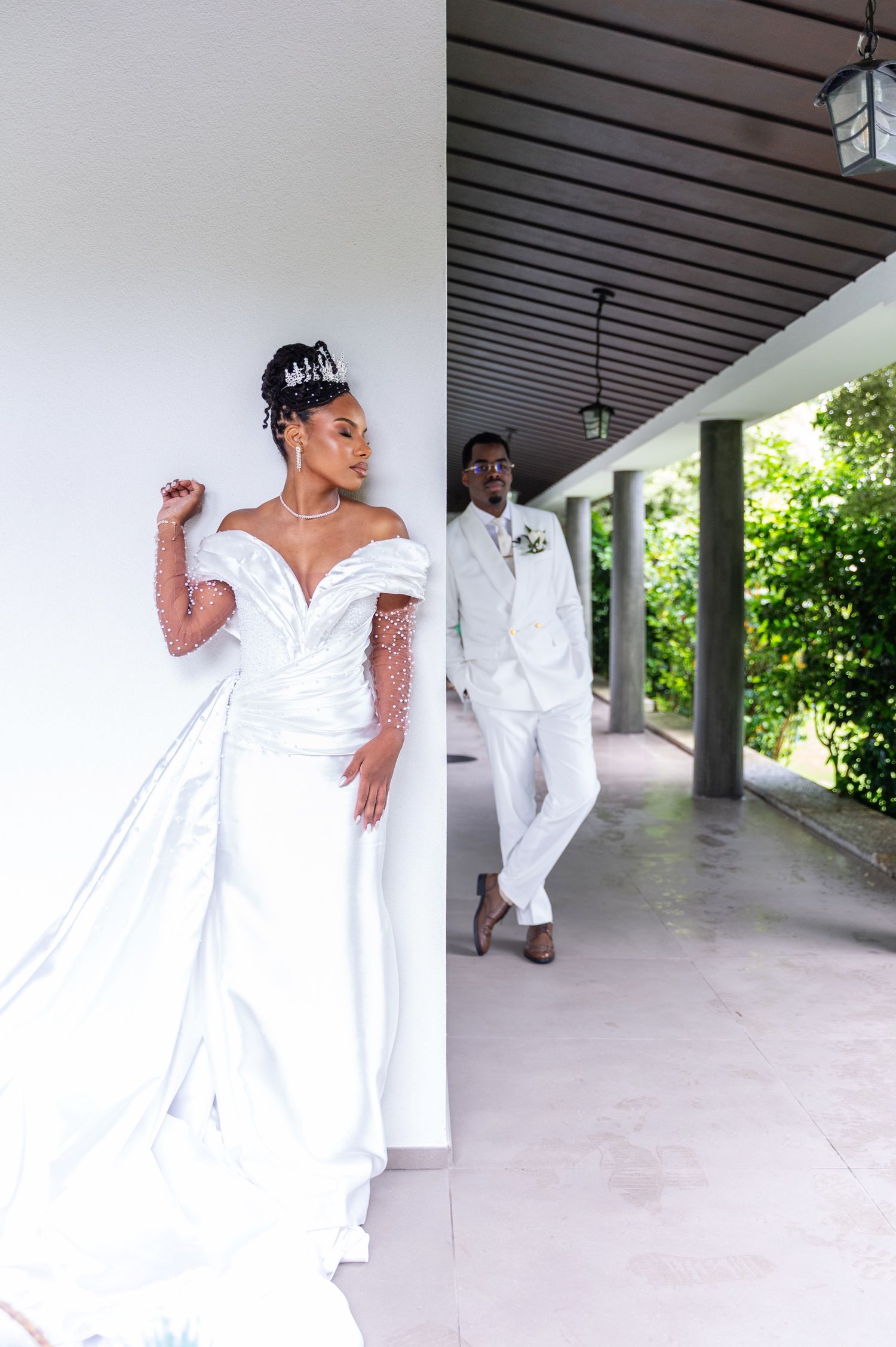 Stunning bride in white pearl gown and crown poses by column while groom stands in background.