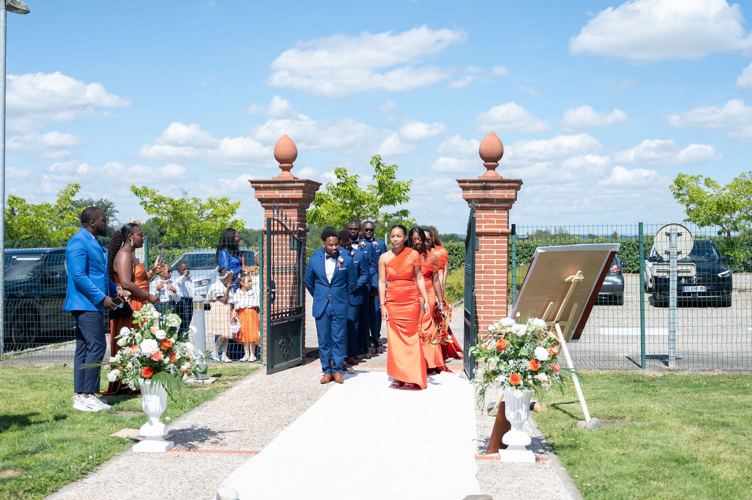 Wedding procession walking down outdoor aisle through brick gates with floral arrangements on sunny day.