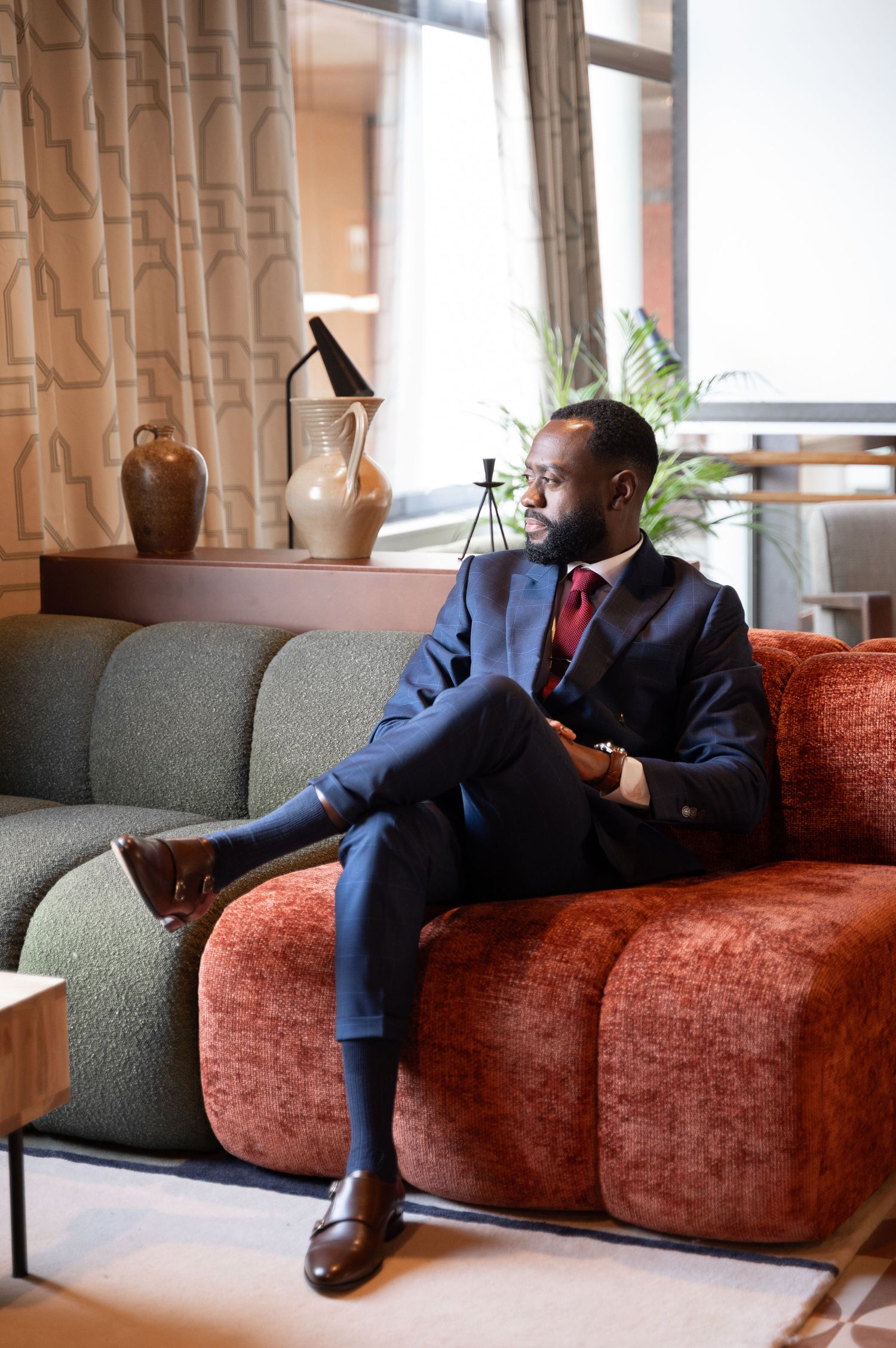 Stylish Black man in navy suit and red tie sits relaxed on a rust-colored sofa in a modern lounge.