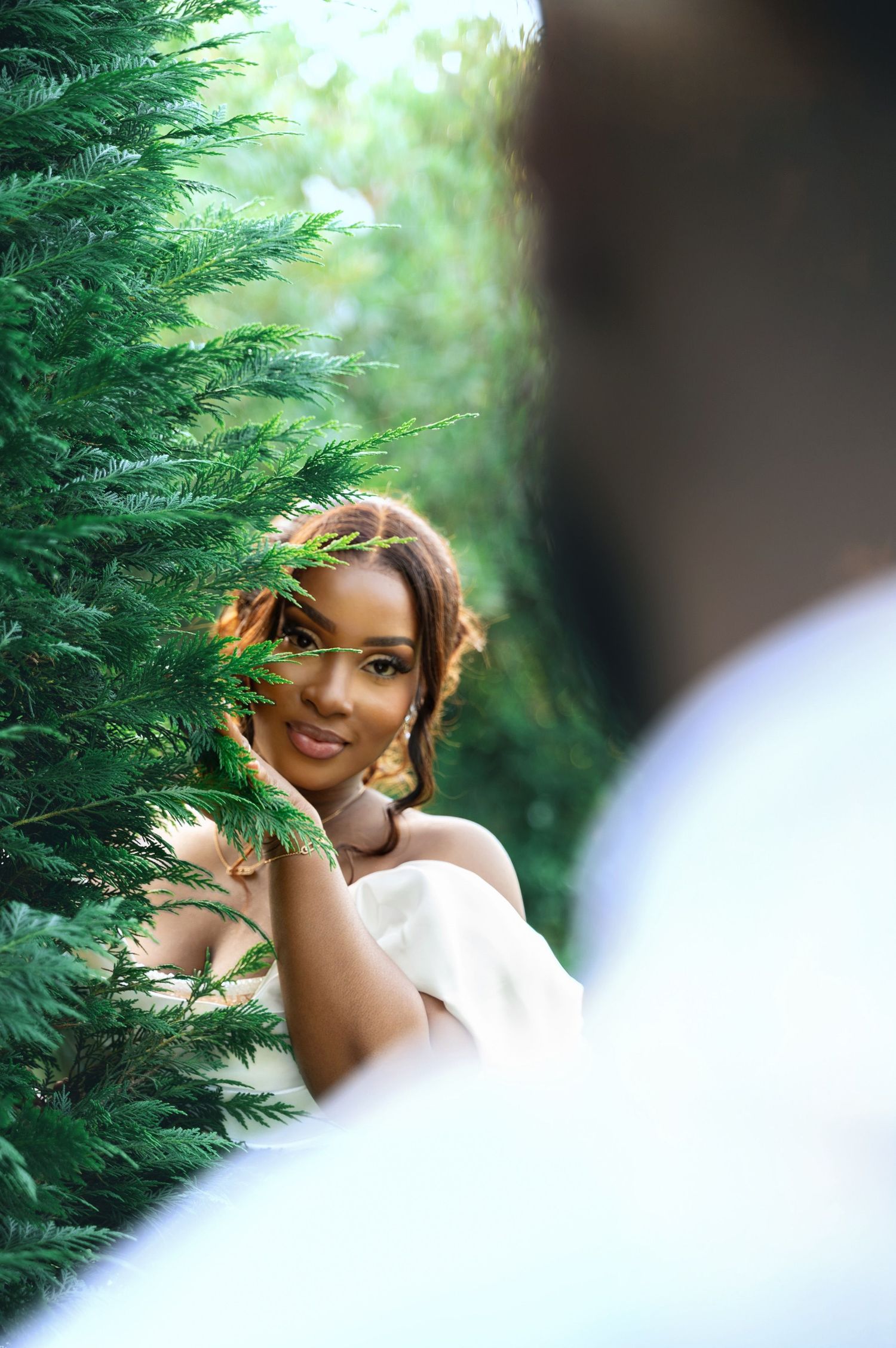 Beautiful Black bride in white gown peeking through lush green cypress trees with a gentle smile.
