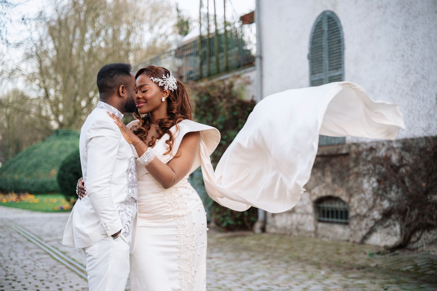Newlywed couple embraces outdoors as bride's veil billows in the wind near a stone building.