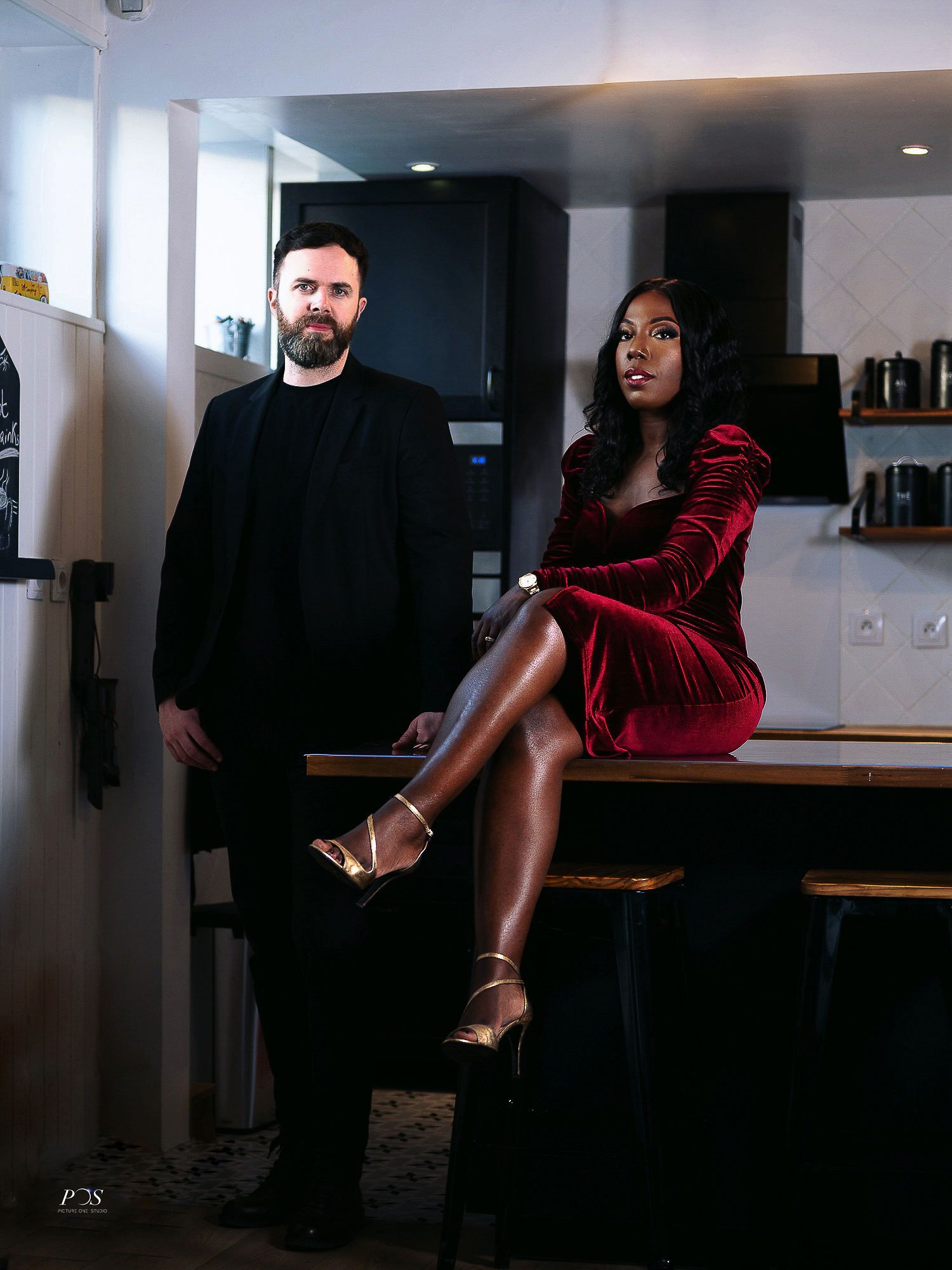 Couple posing in a modern kitchen, man in black suit and woman in red velvet dress with gold heels.