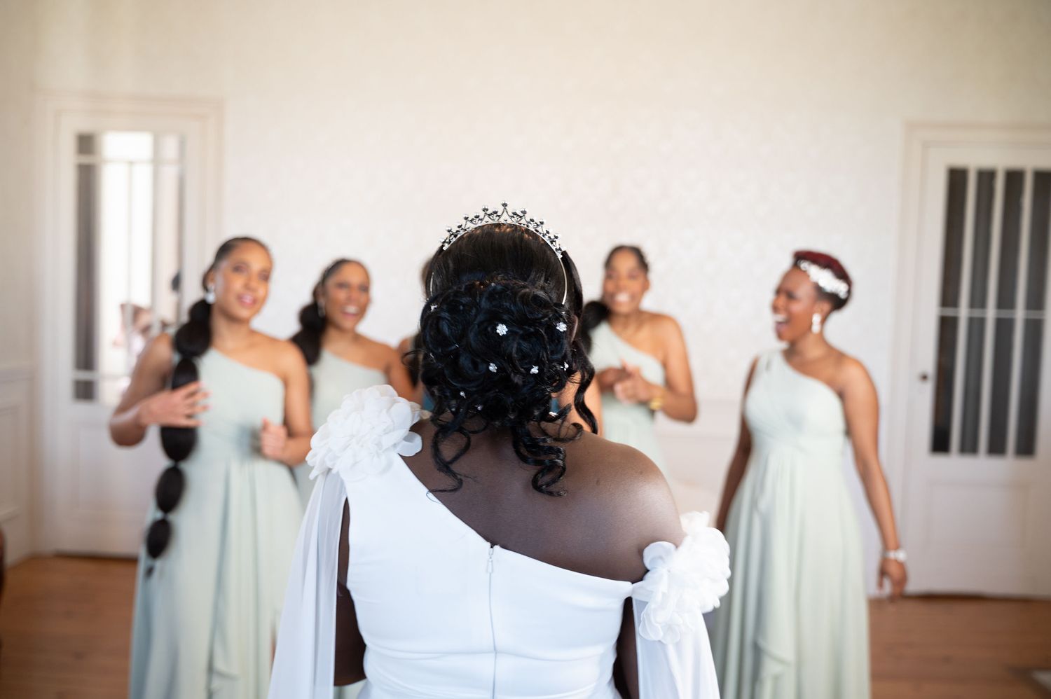 Bride in white gown with pearl hair accessories faces smiling bridesmaids in sage green dresses during wedding reveal.