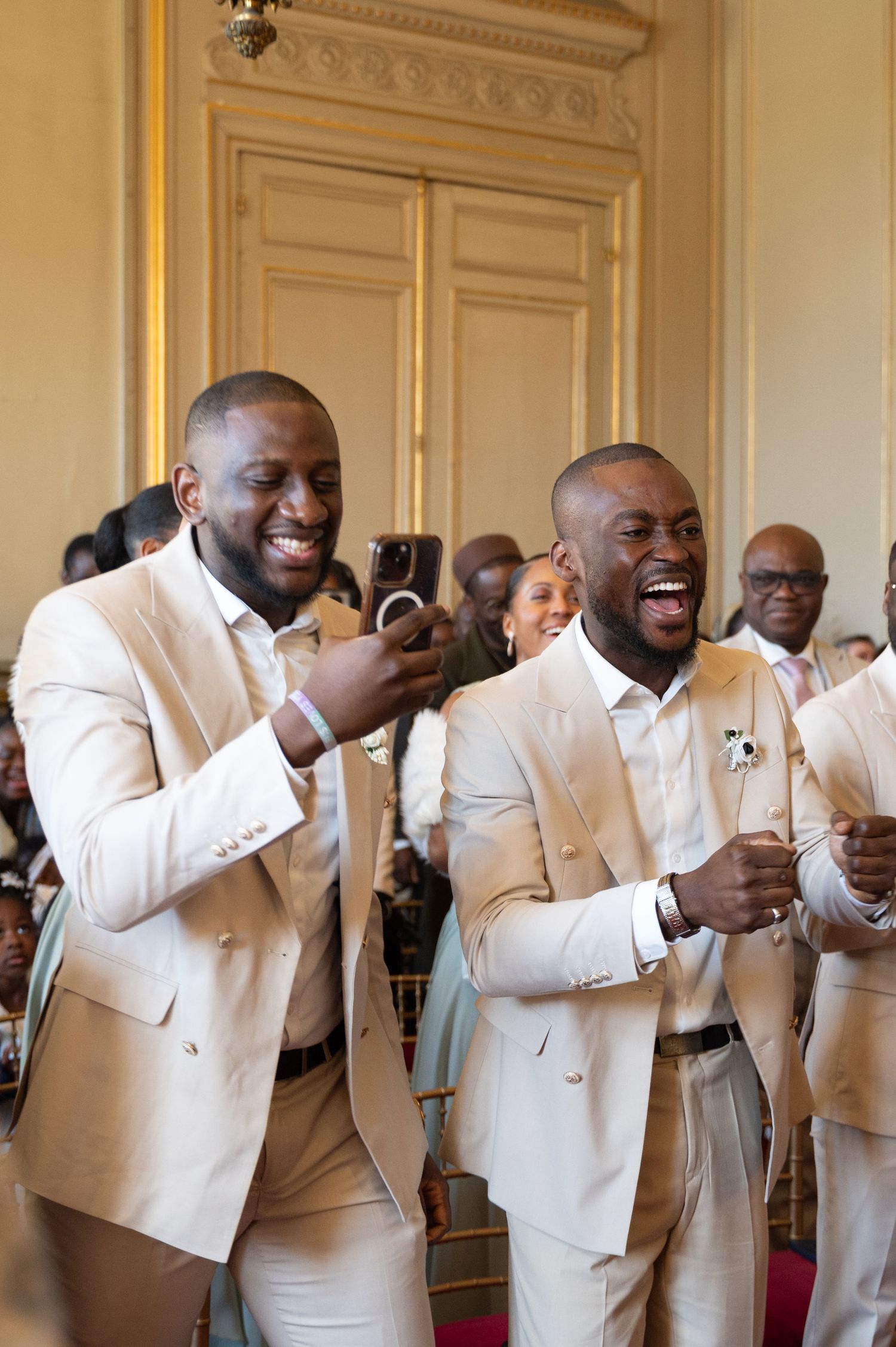 Two men in beige suits laughing joyfully during wedding ceremony indoors.