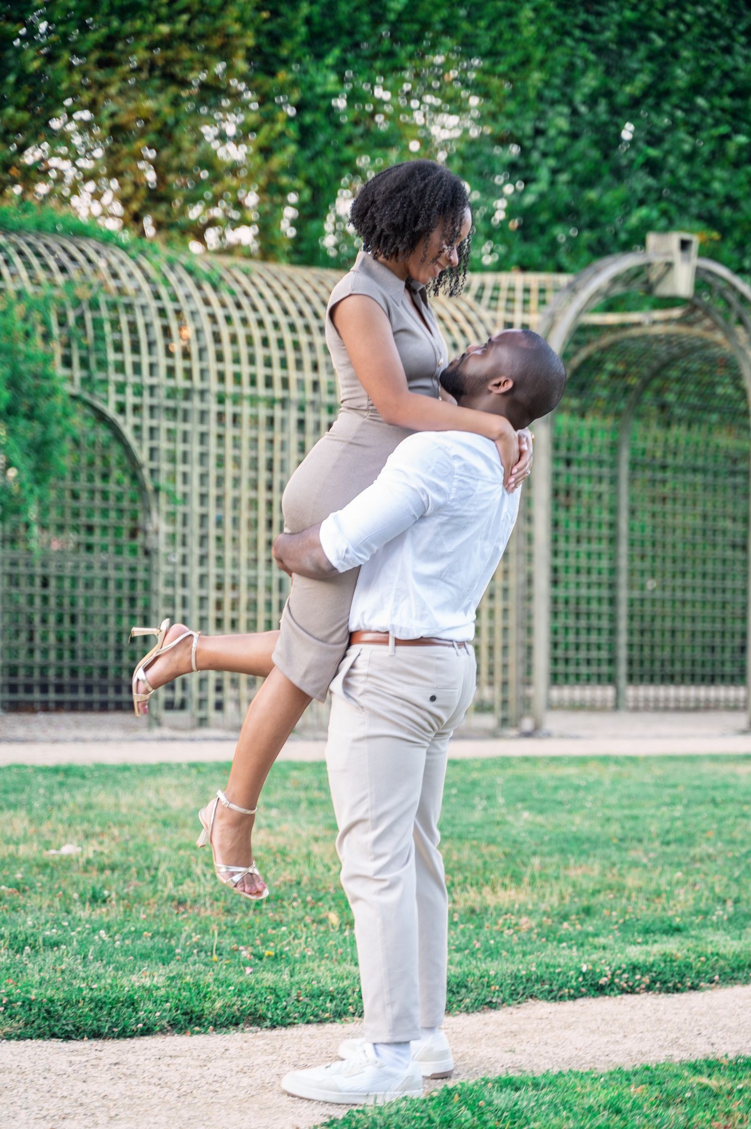 A happy Black couple shares a playful lift and laugh together in front of a wooden trellis garden wall.