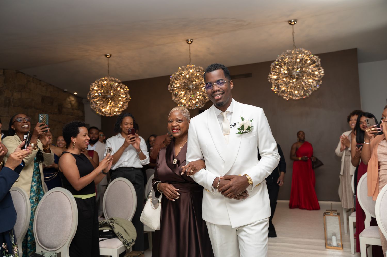 Groom in white suit walks into wedding reception escorted by a woman under golden chandelier lights.