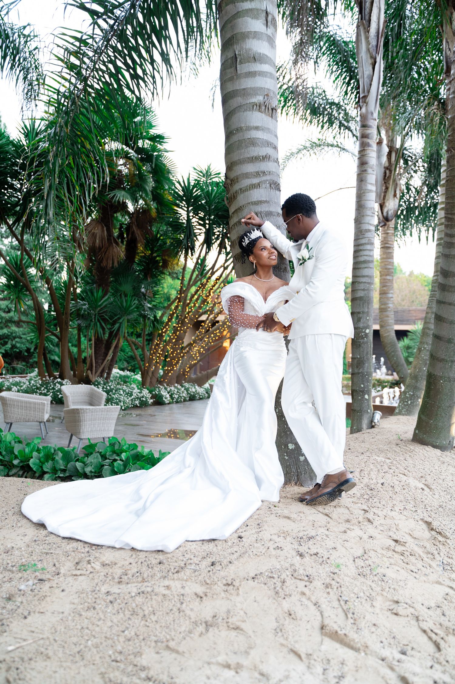 Groom in white suit adjusts brides crystal crown as she sits in flowing gown on sandy ground.