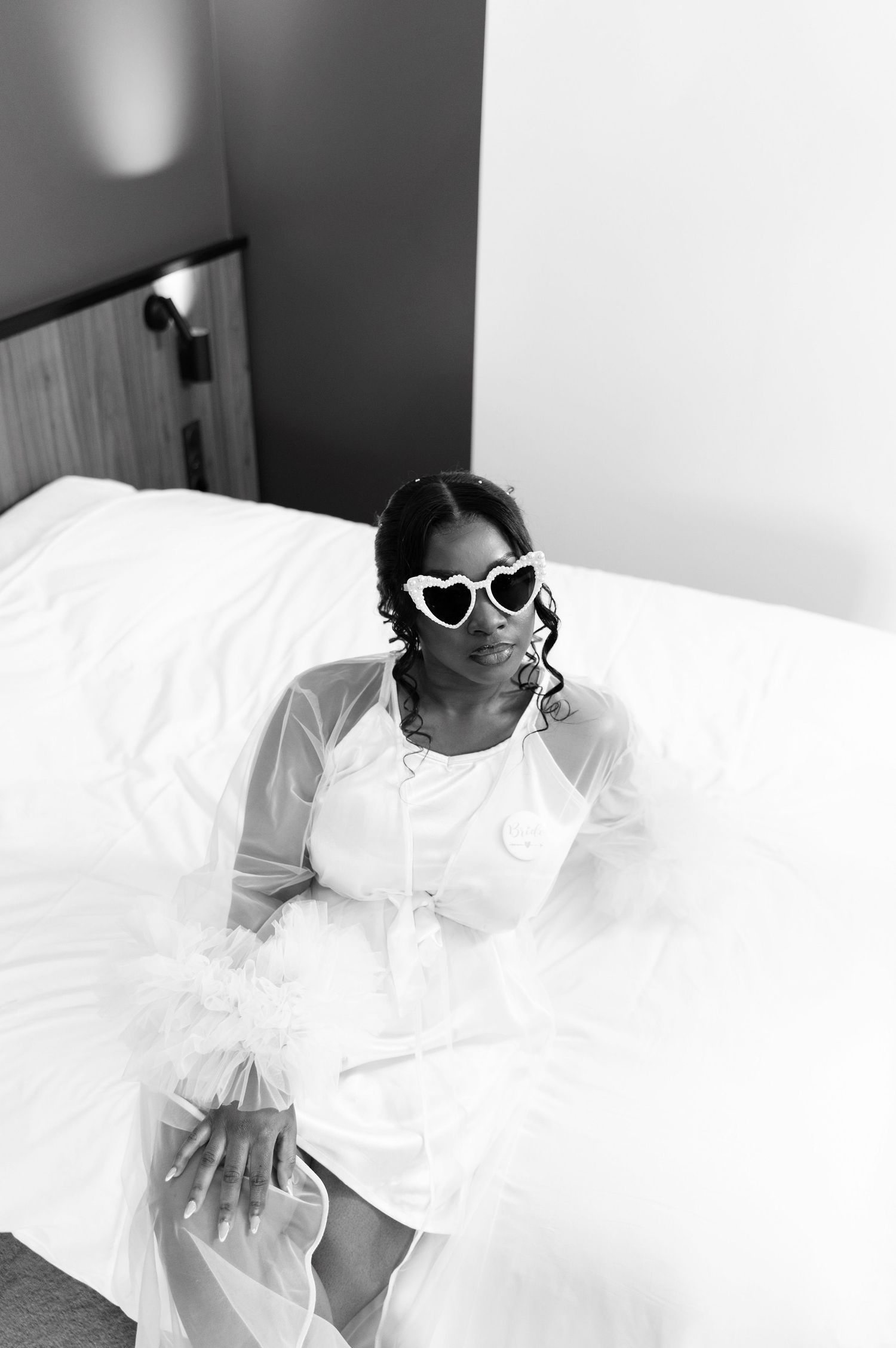 Black and white photo of a bride in white robe and heart-shaped sunglasses sitting on a hotel bed.