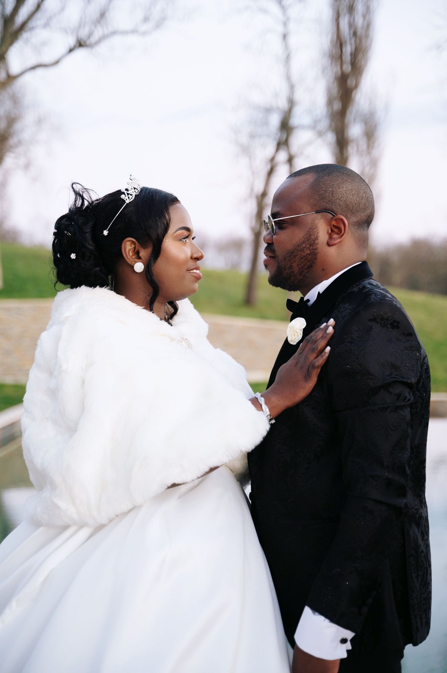 Bride in white fur wrap and groom in tuxedo sharing intimate moment outdoors.