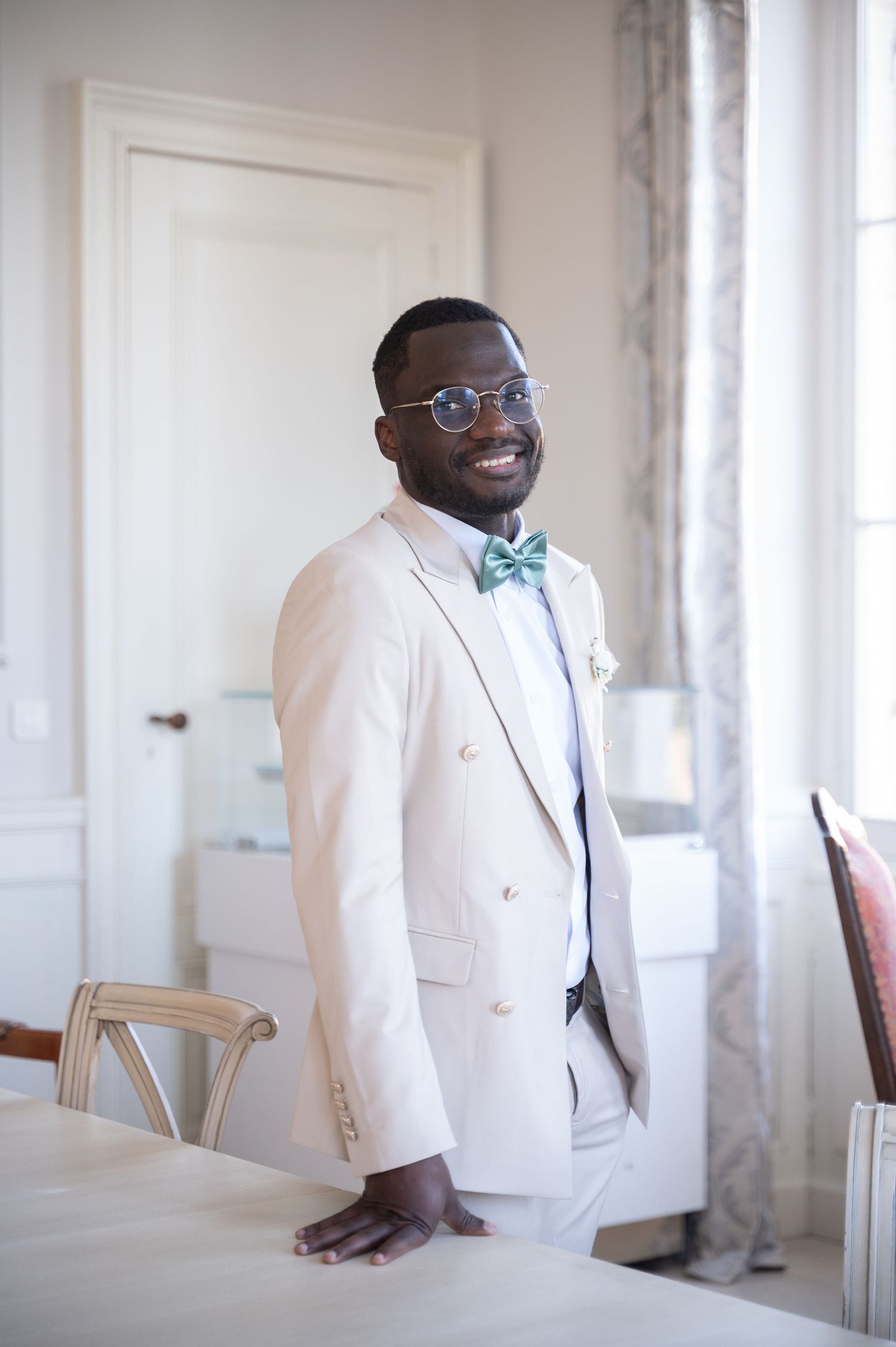 Smiling groom in cream double-breasted suit with green bow tie and glasses at elegant wedding venue.