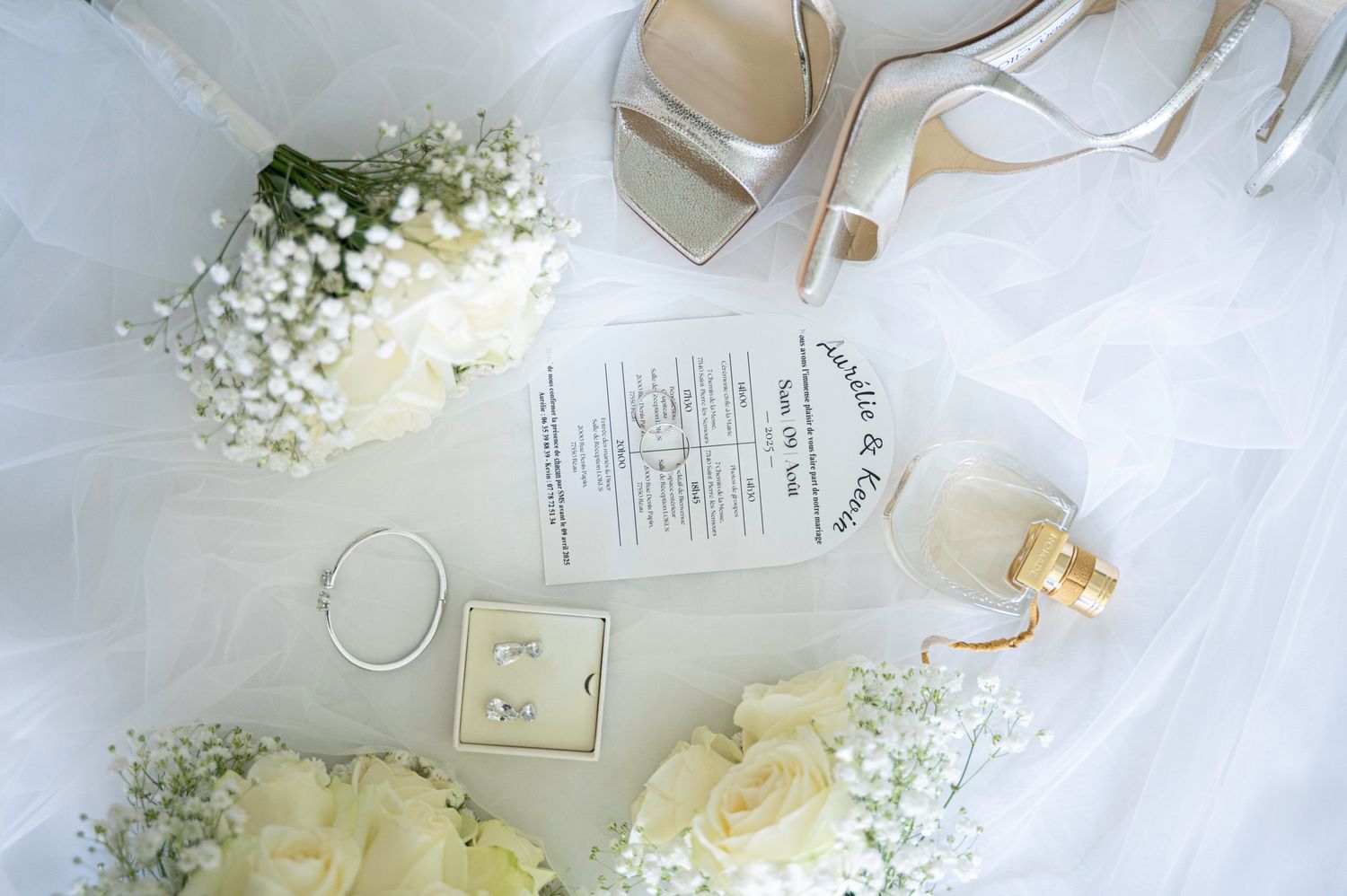 Bridal flat lay with white roses, Jimmy Choo heels, wedding rings, and ceremony program on veil.