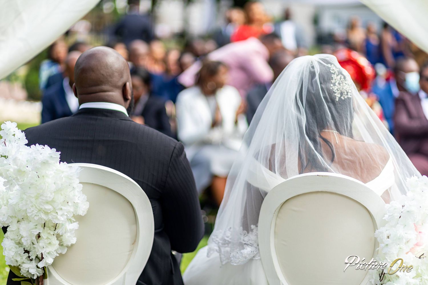 Bride in veil and groom in tuxedo seated at outdoor wedding ceremony with white floral arrangements.