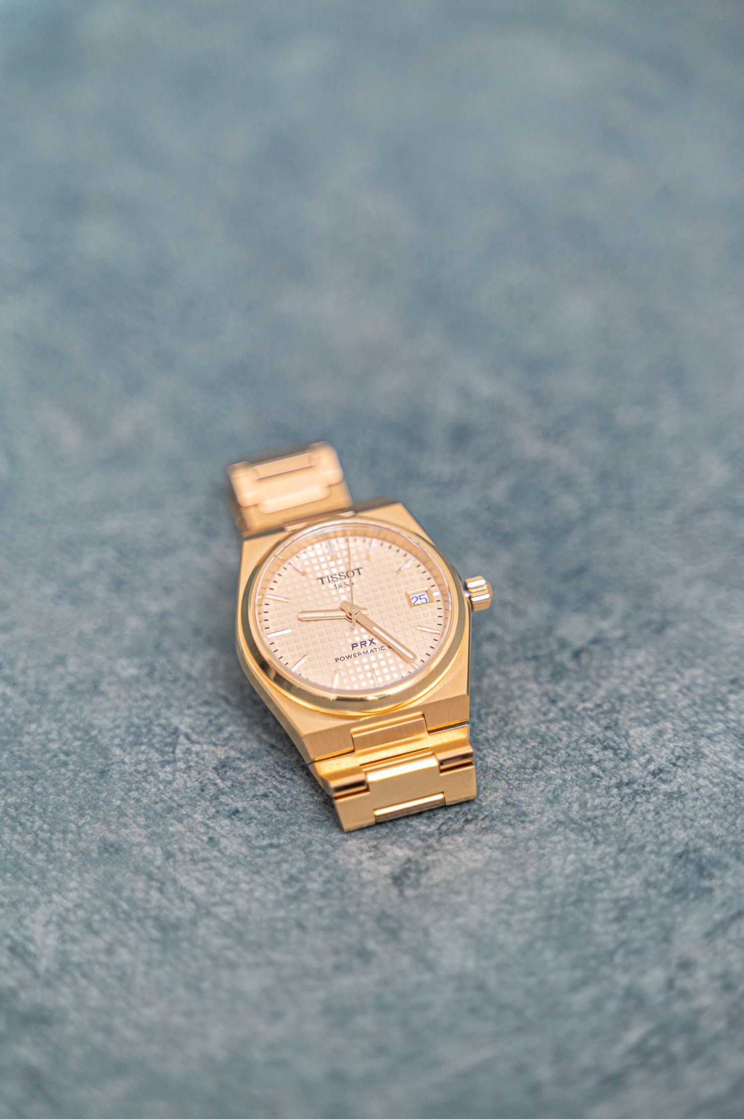 Gold Tissot PRX watch with rose gold dial on blue-gray textured surface.