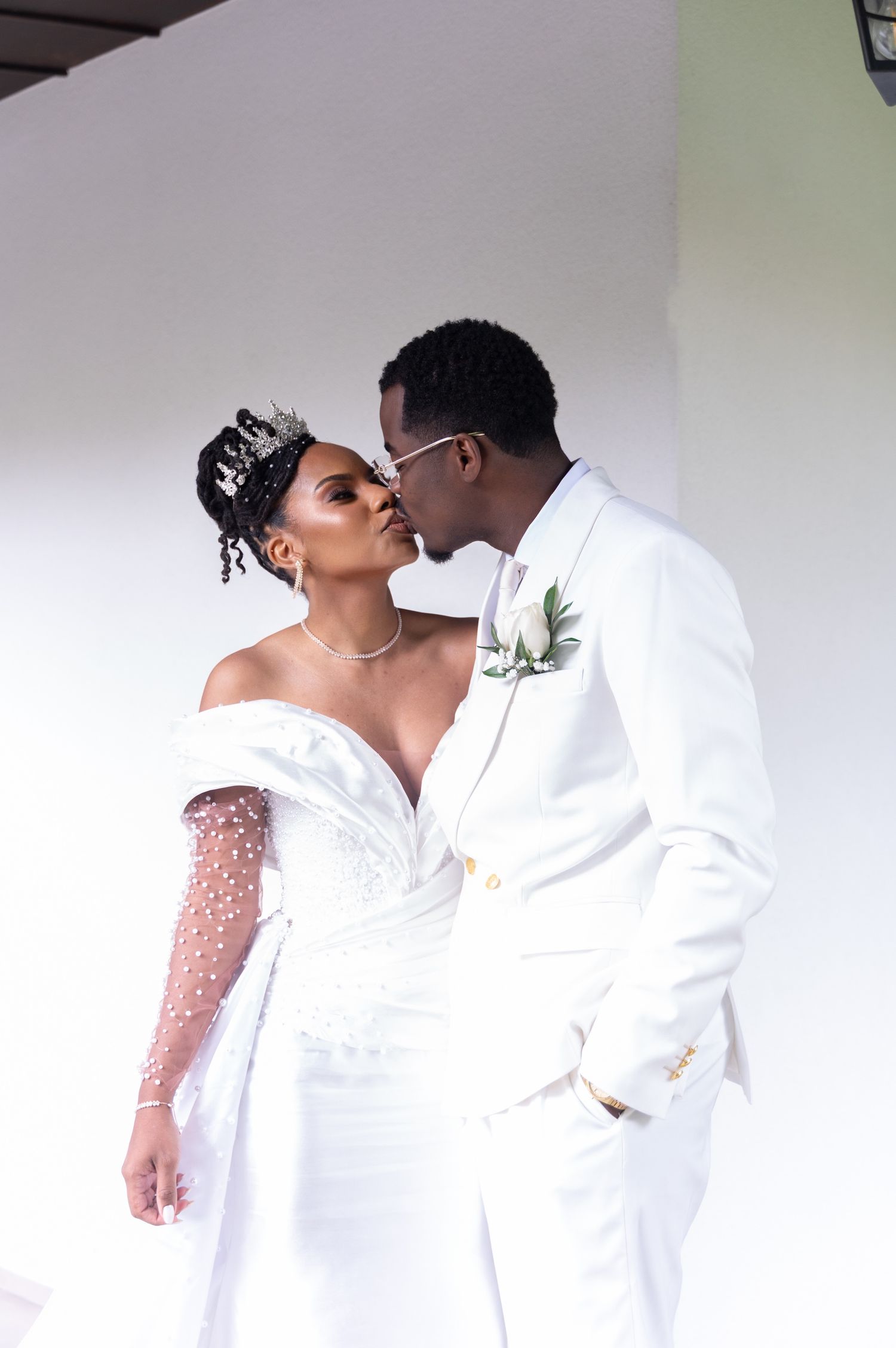 Black bride and groom sharing romantic kiss in matching white wedding attire with floral boutonniere.
