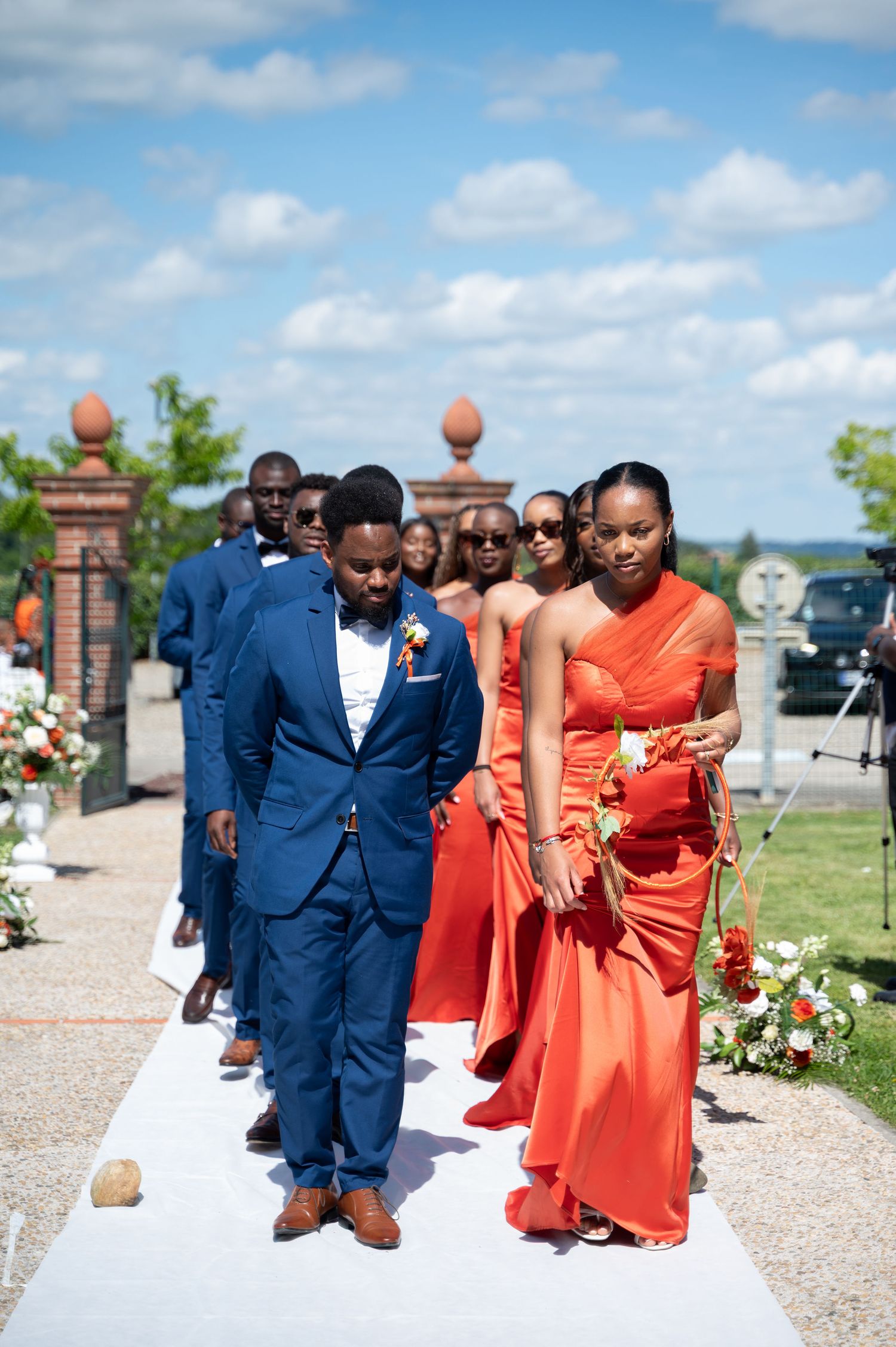 Groomsmen in blue suits and bridesmaids in orange dresses walk down wedding aisle outdoors.