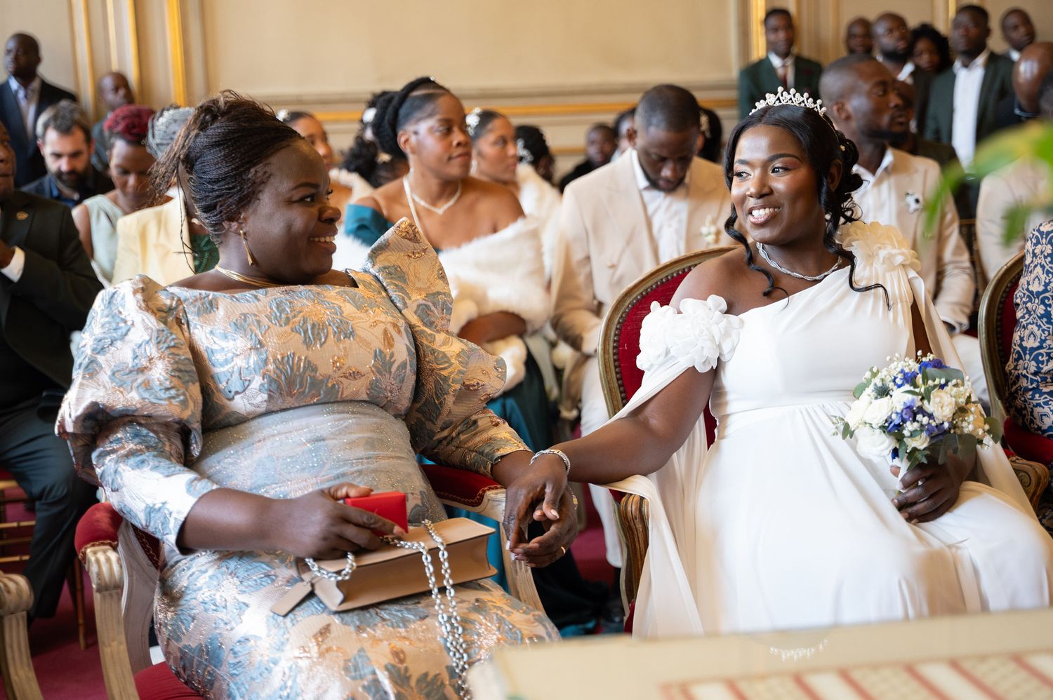 Bride in white gown holding bouquet shares tender moment with seated elder at wedding.