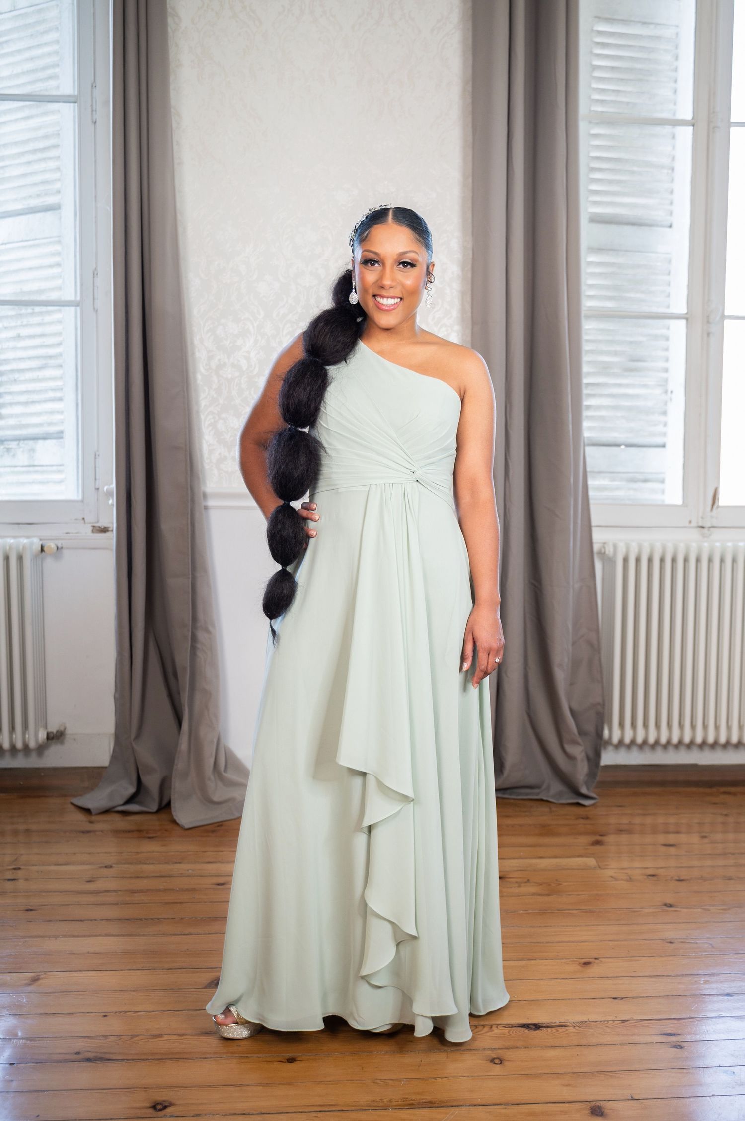 Woman in sage green one-shoulder bridesmaid gown with long braided ponytail posing in bright room.