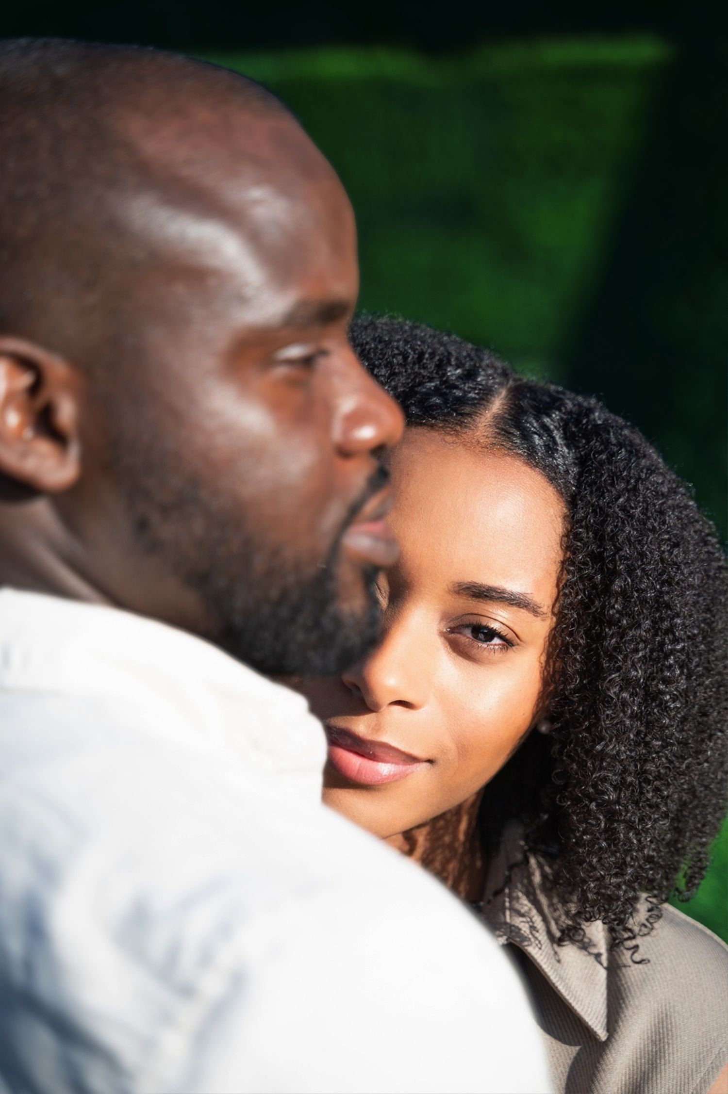 A man tenderly kisses a womans temple as she gazes softly forward against a lush green background.