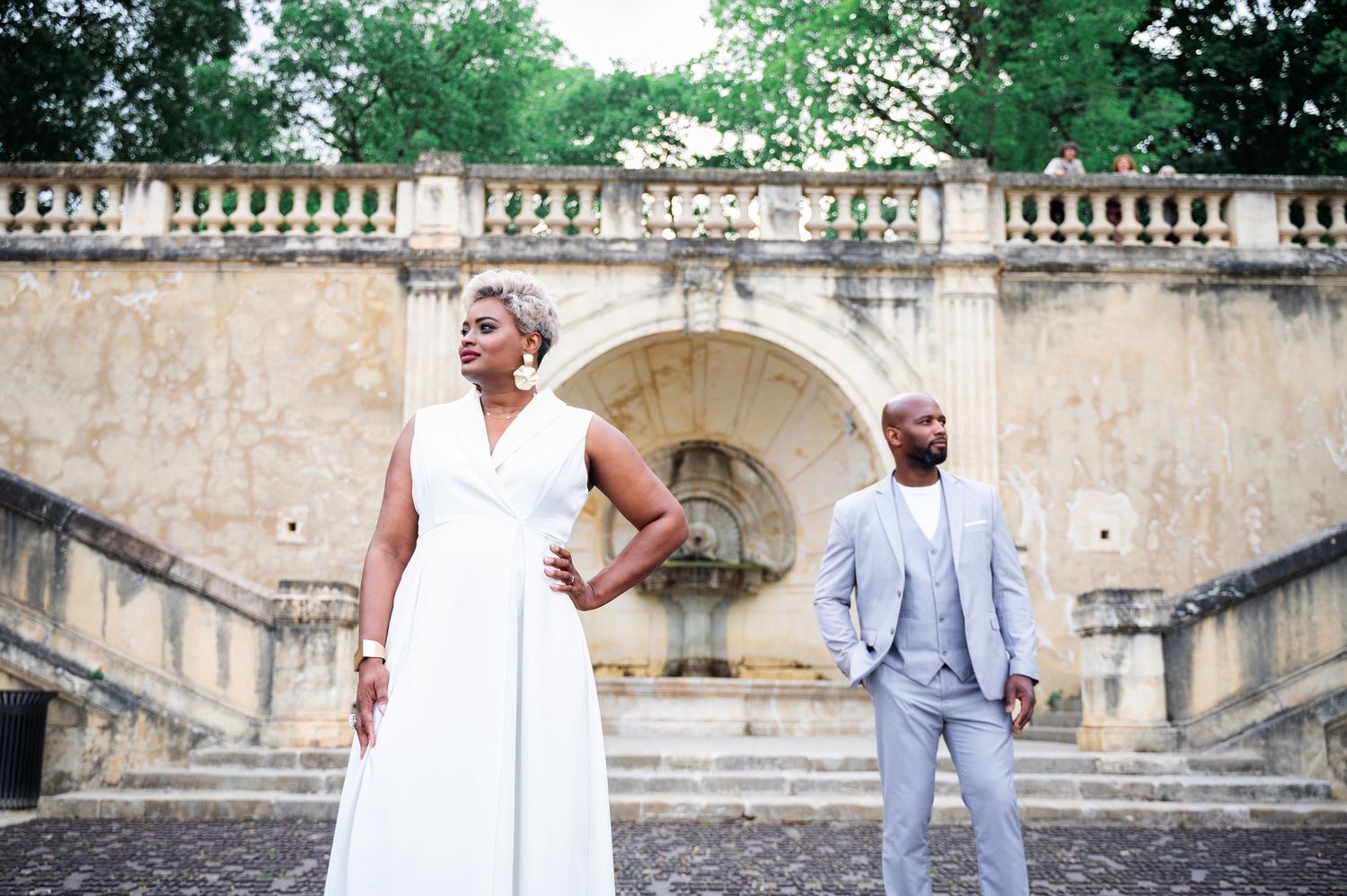 Elegant Black couple posing at ornate stone fountain, woman in white dress, man in light gray suit.