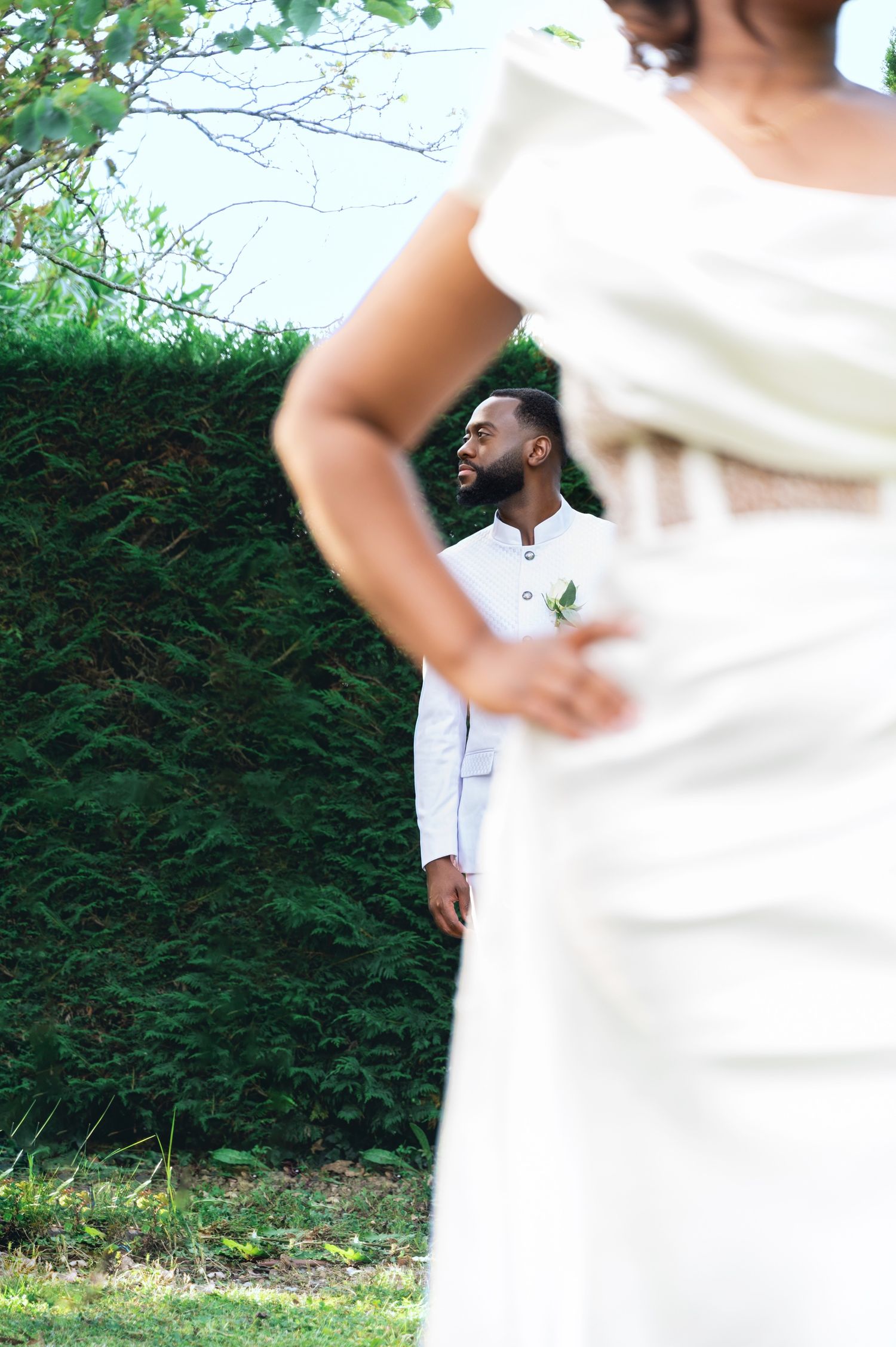 A groom in white attire smiles as he catches his first glimpse of his bride in a lush green garden setting.