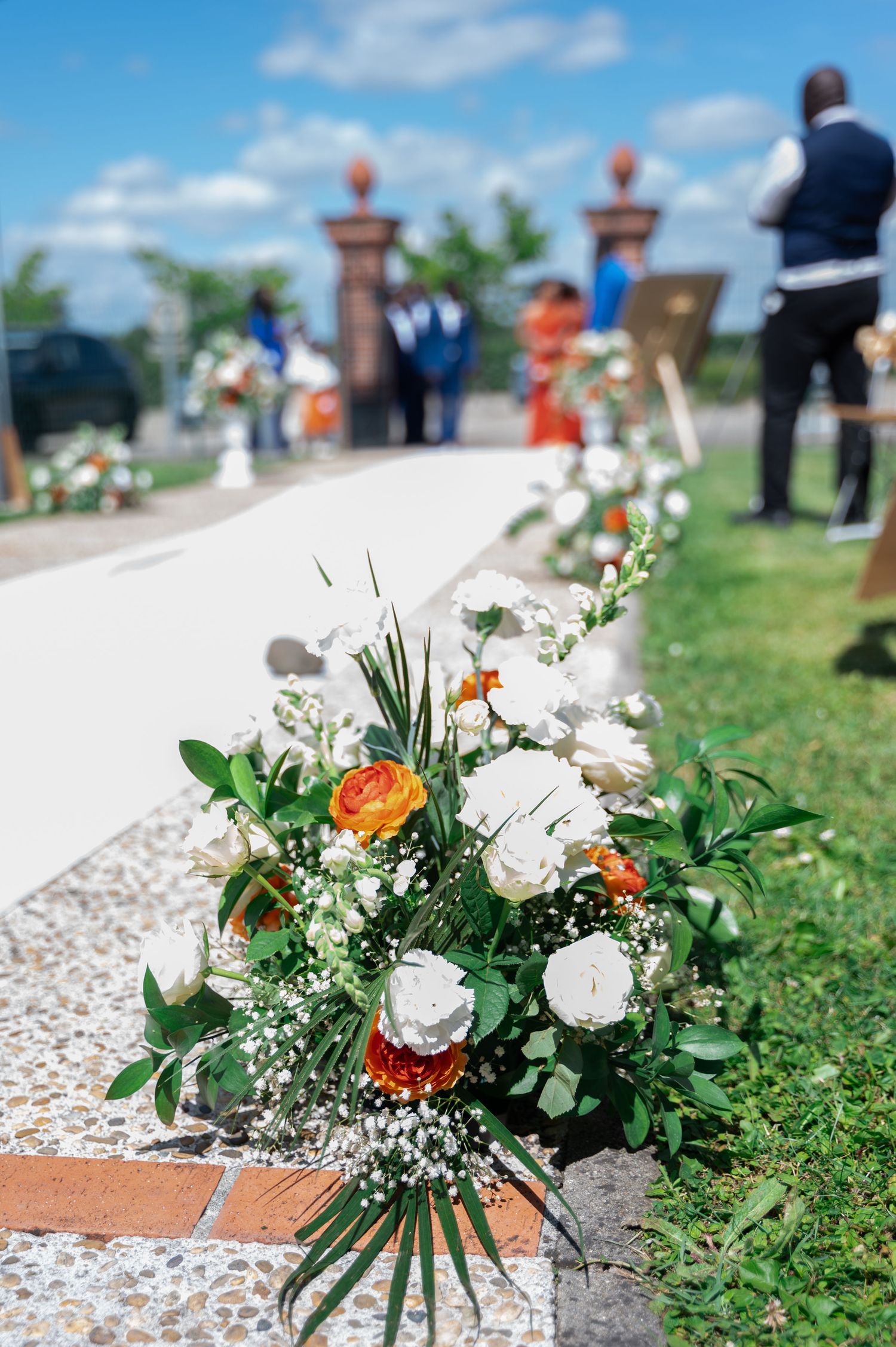 White and orange floral arrangements line the wedding aisle path at sunny outdoor ceremony venue.