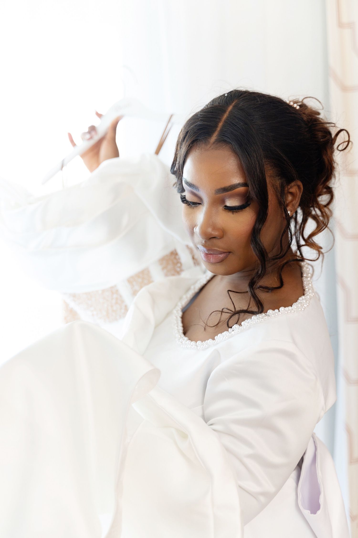 Radiant bride in white robe with delicate lace details posing near bright window.