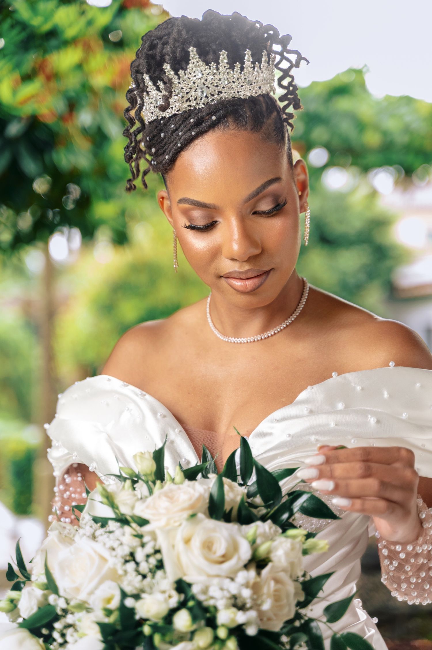 Black bride in tiara and pearl wedding gown holds white rose bouquet outdoors.