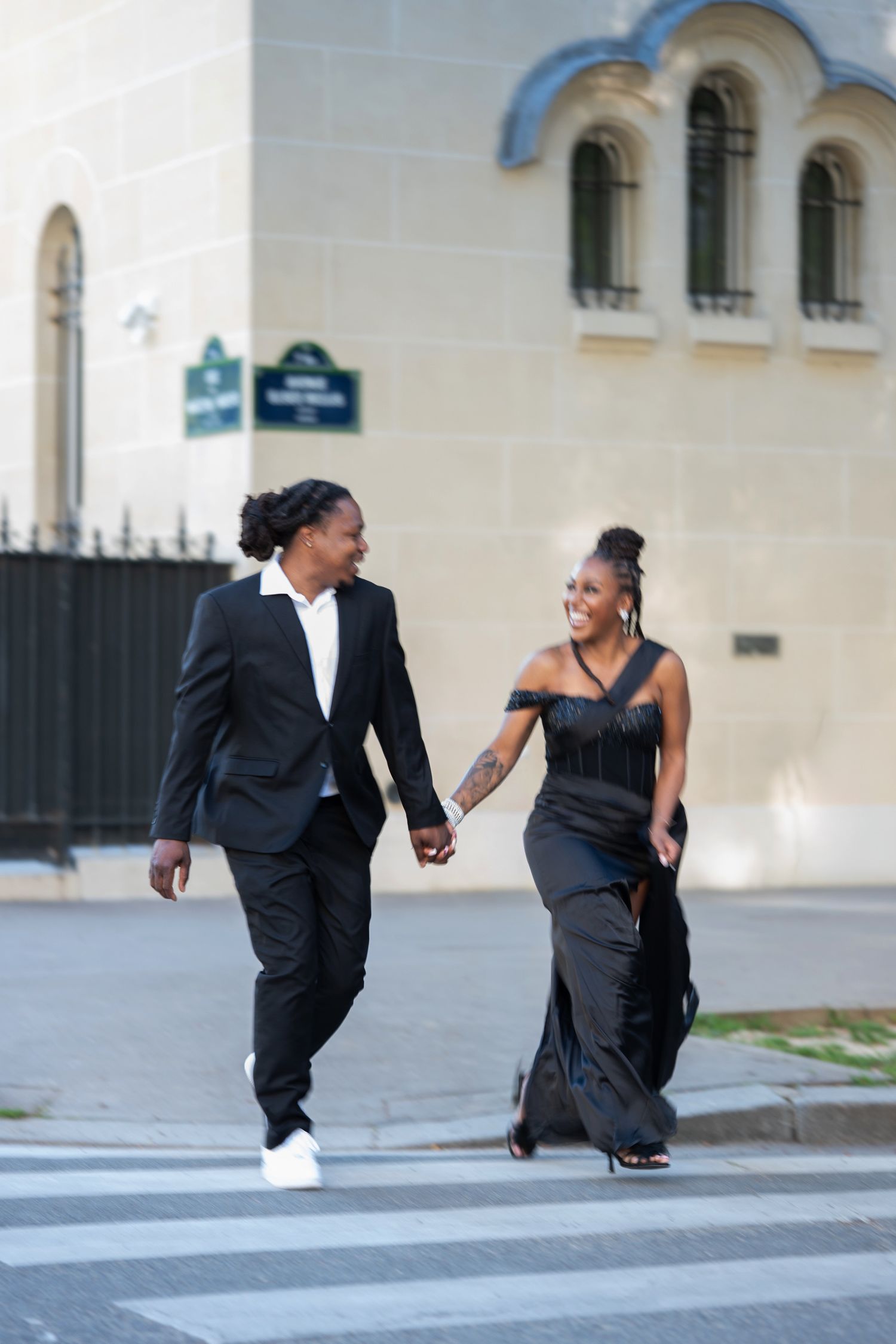 Stylish Black couple holding hands crossing a street in Paris, dressed in elegant black outfits, smiling at each other.