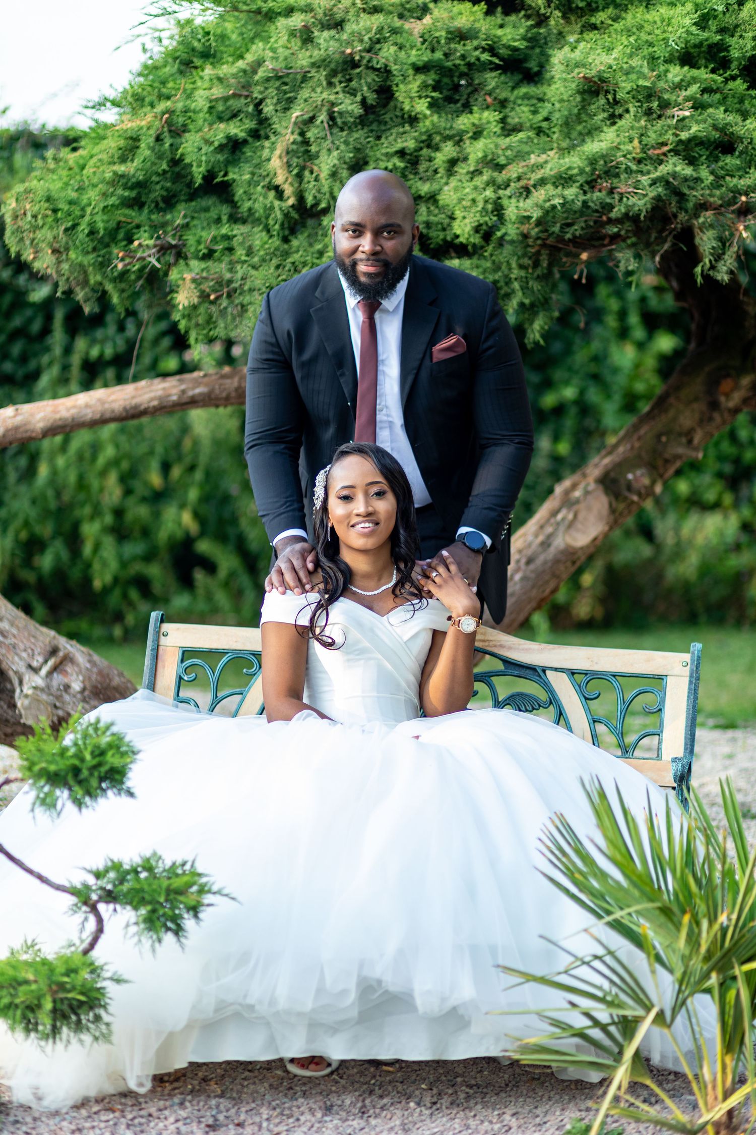 Bride in white gown sits on garden bench while groom in black suit stands behind her outdoors.
