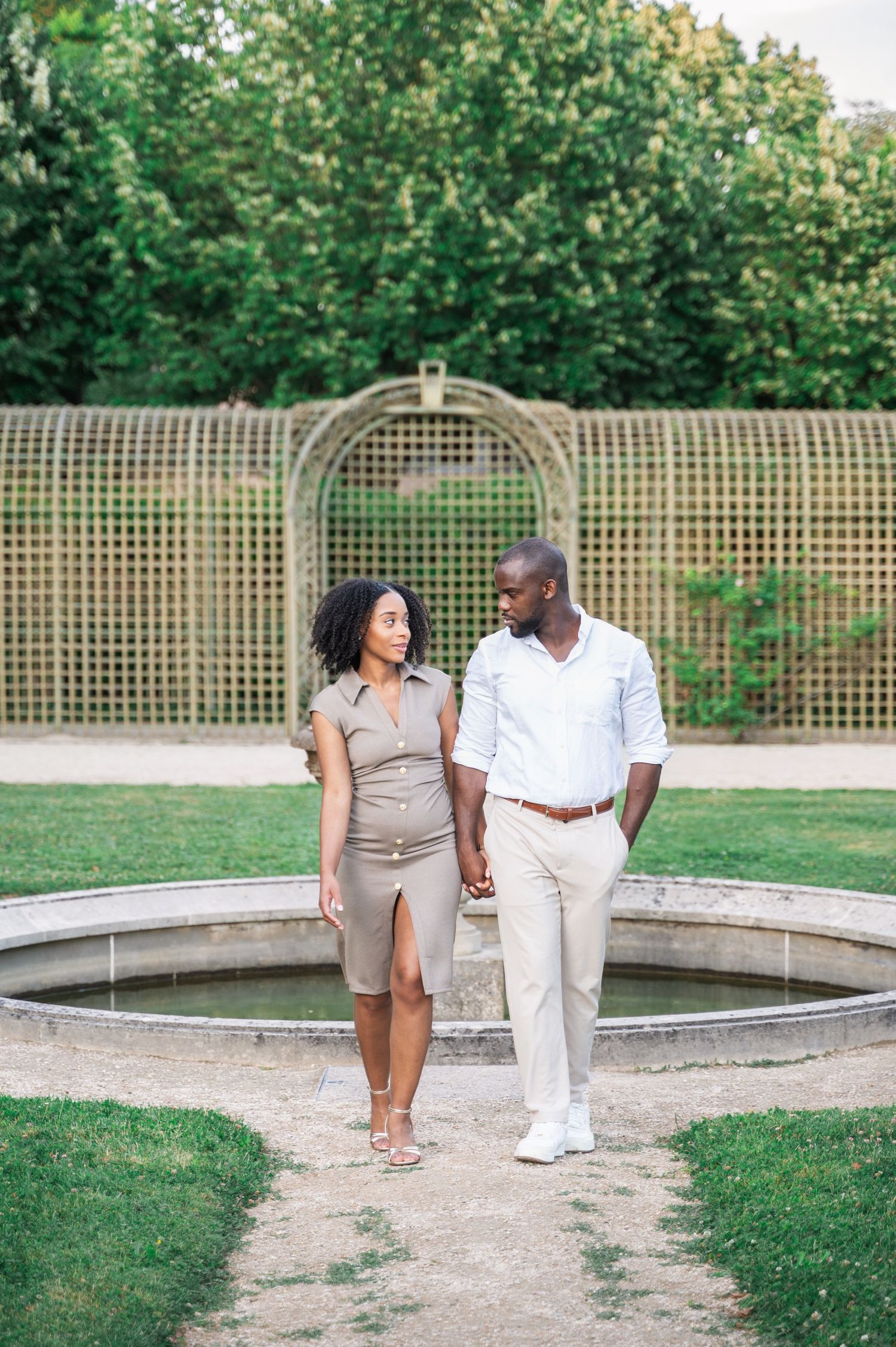 A stylish Black couple holds hands and gazes at each other while walking near a circular fountain in a park.