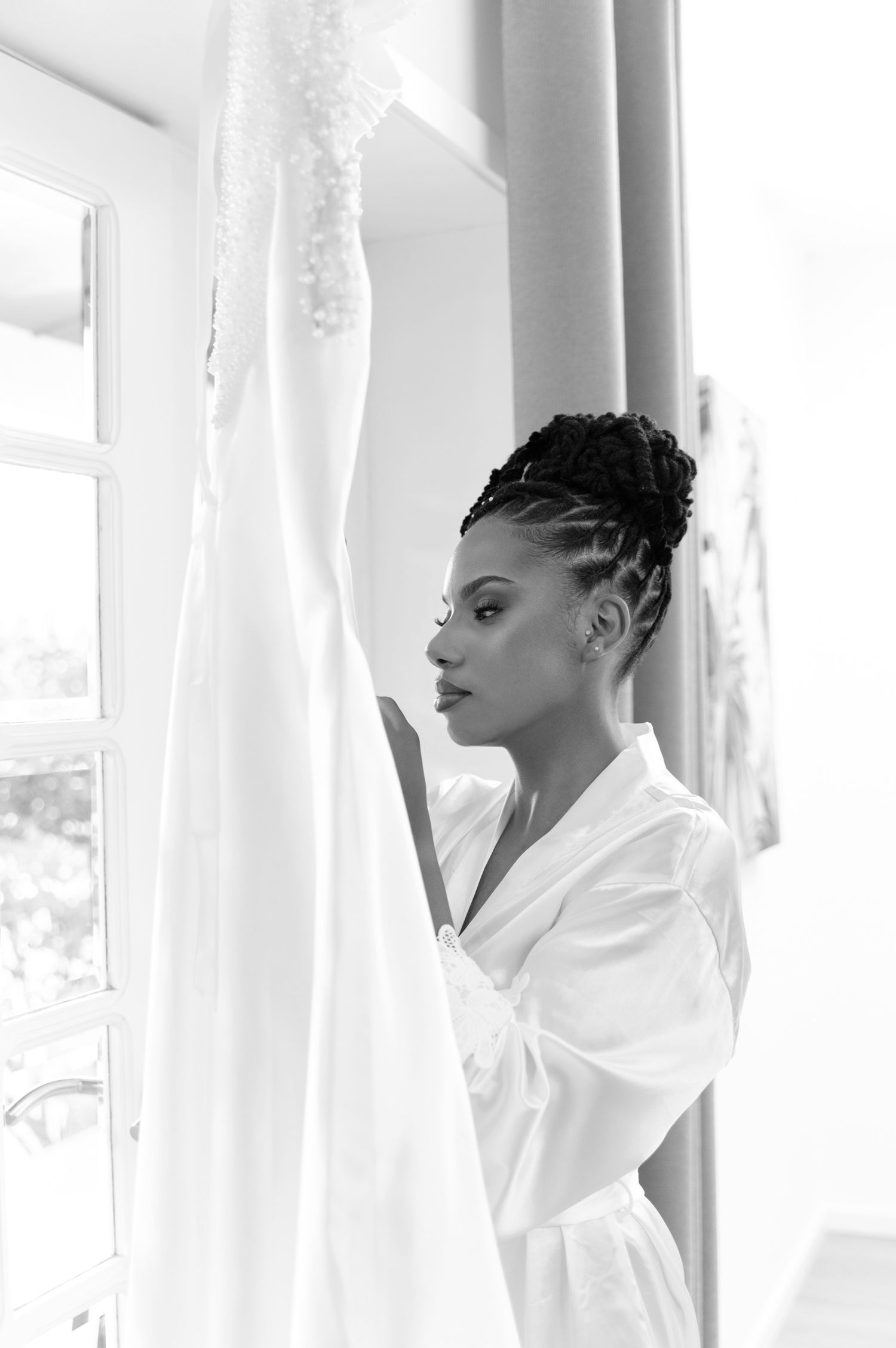 Black and white portrait of bride in white robe gazing at her hanging wedding gown.