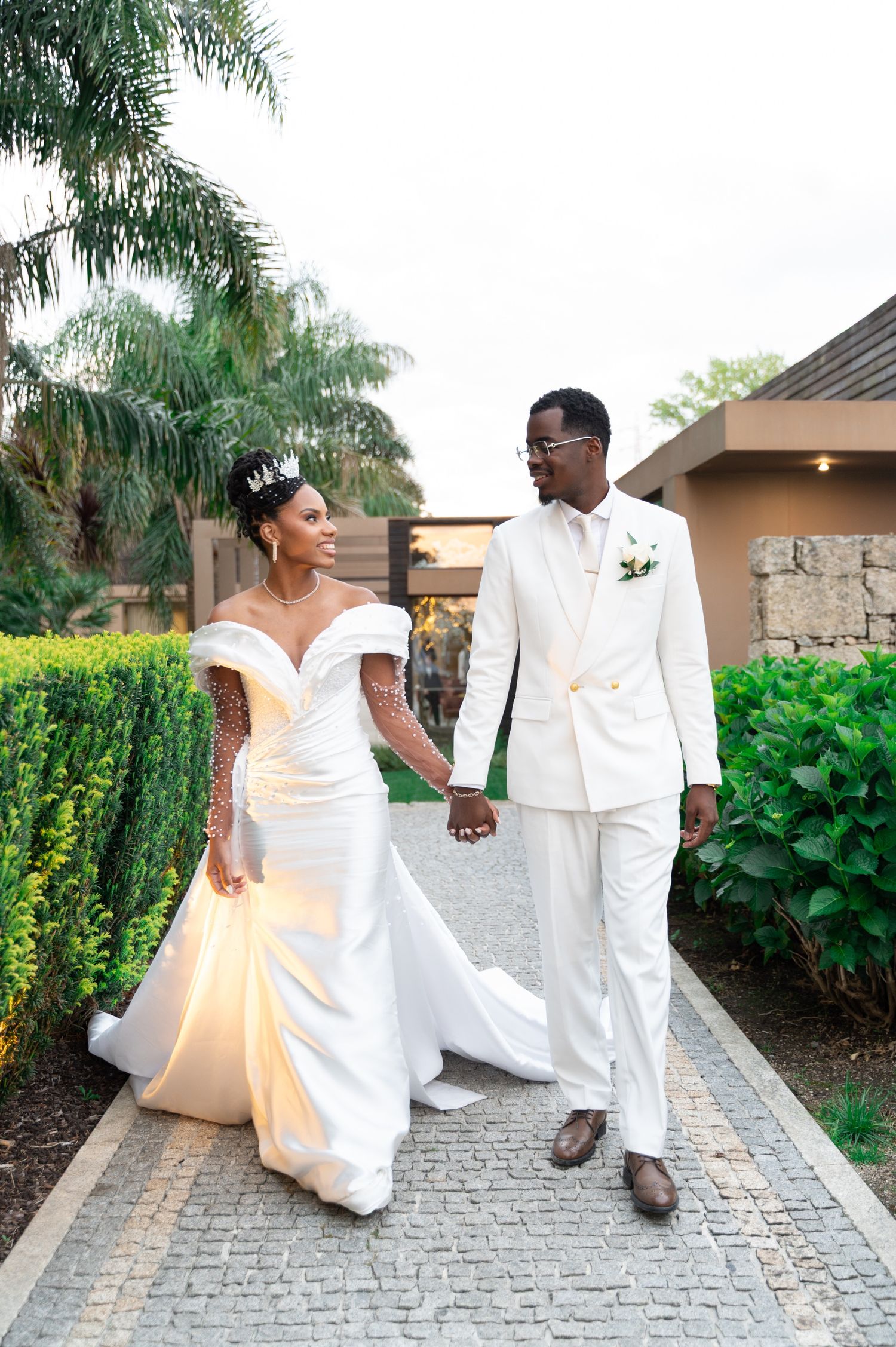 Elegant bride and groom in white wedding attire walking hand in hand on a cobblestone path surrounded by lush greenery.