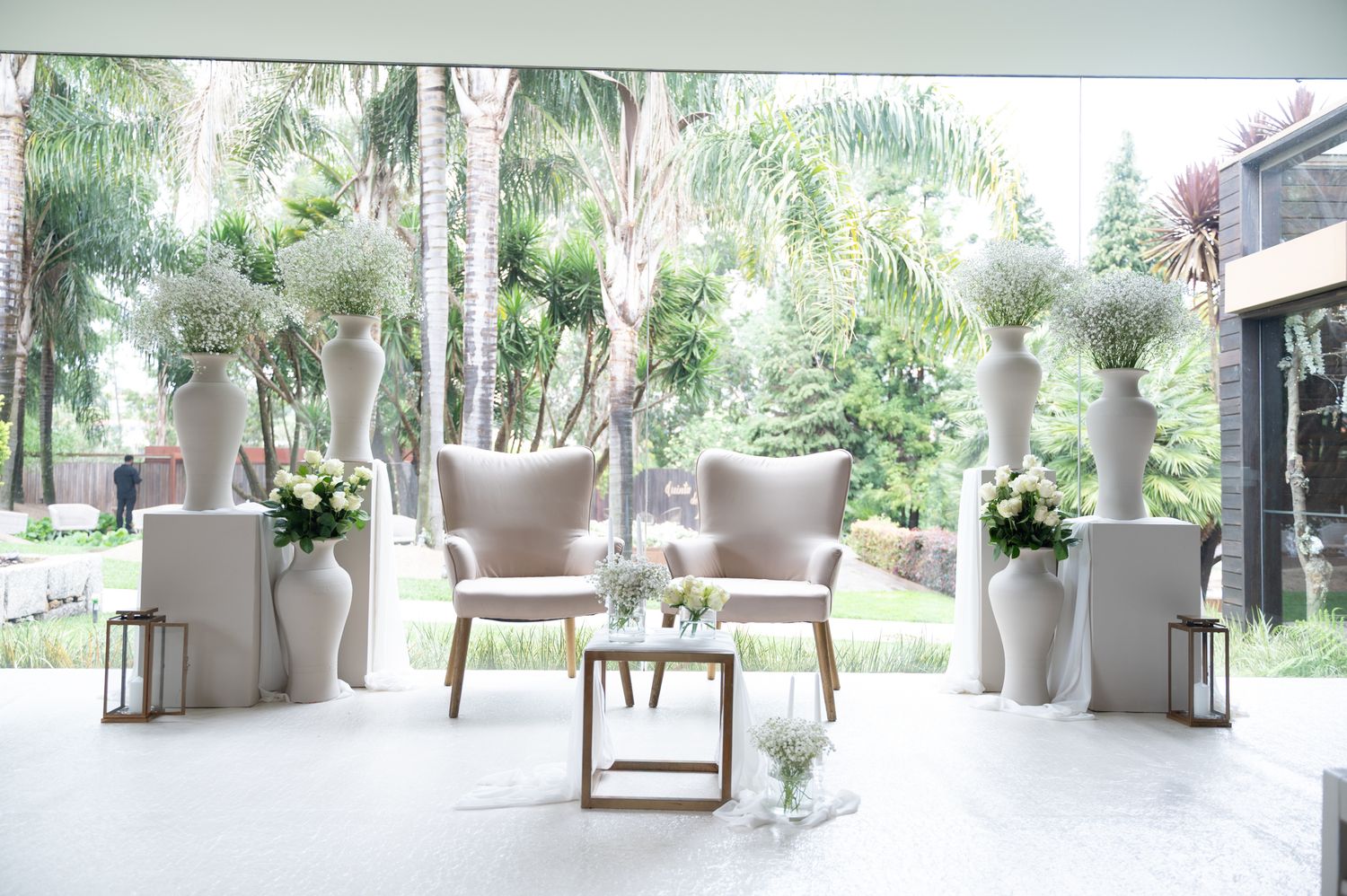 Elegant white wedding ceremony setup with tall vases, floral arrangements, and two chairs in a bright venue.