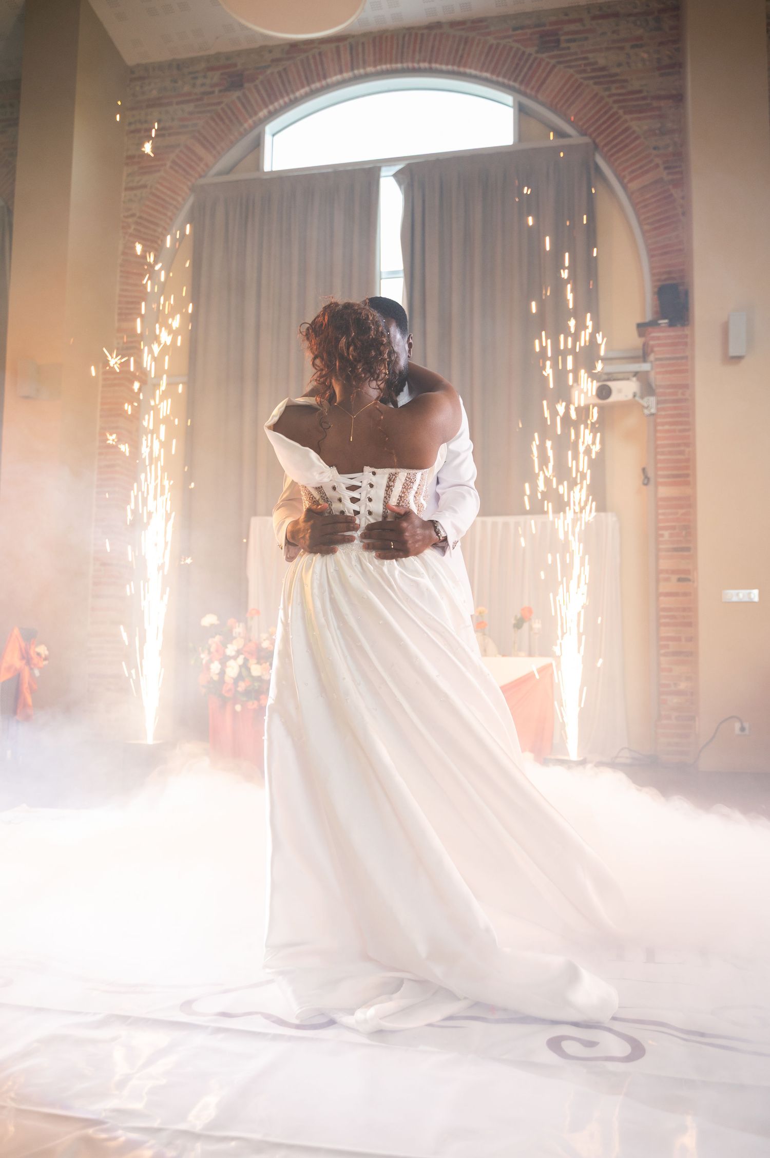 Bride in white off-shoulder gown dancing amid sparkling fireworks and fog at wedding celebration.