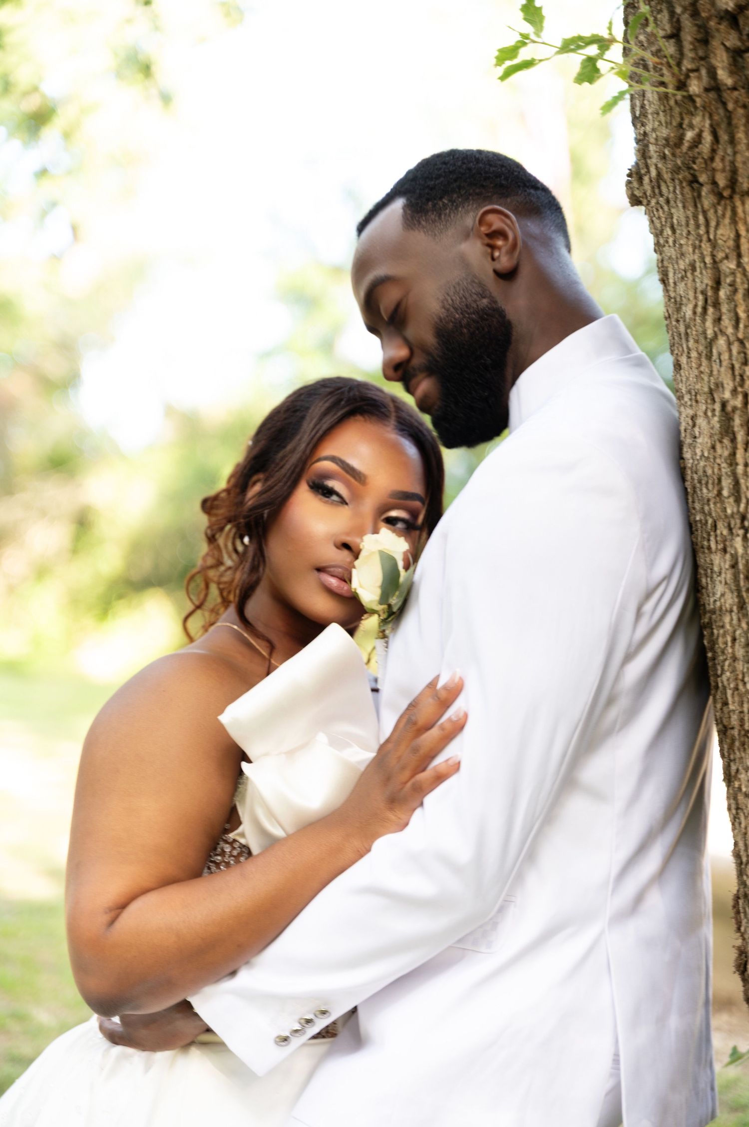 A Black bride in a white gown embraces her groom as they lean against a tree in a romantic pose.