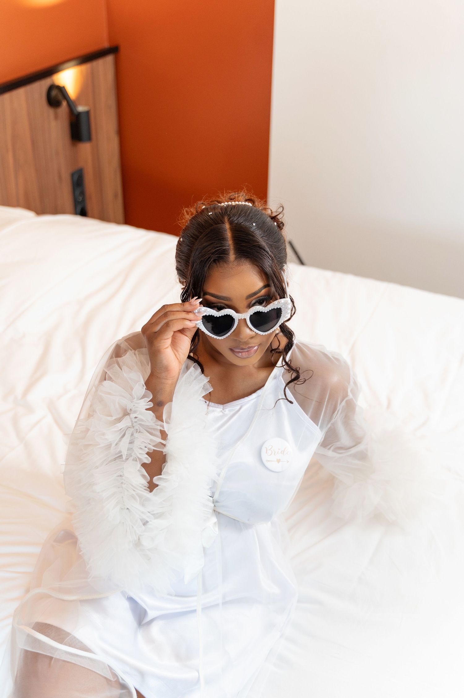 Bride in white feather robe adjusting heart-shaped sunglasses while sitting on a white bed.