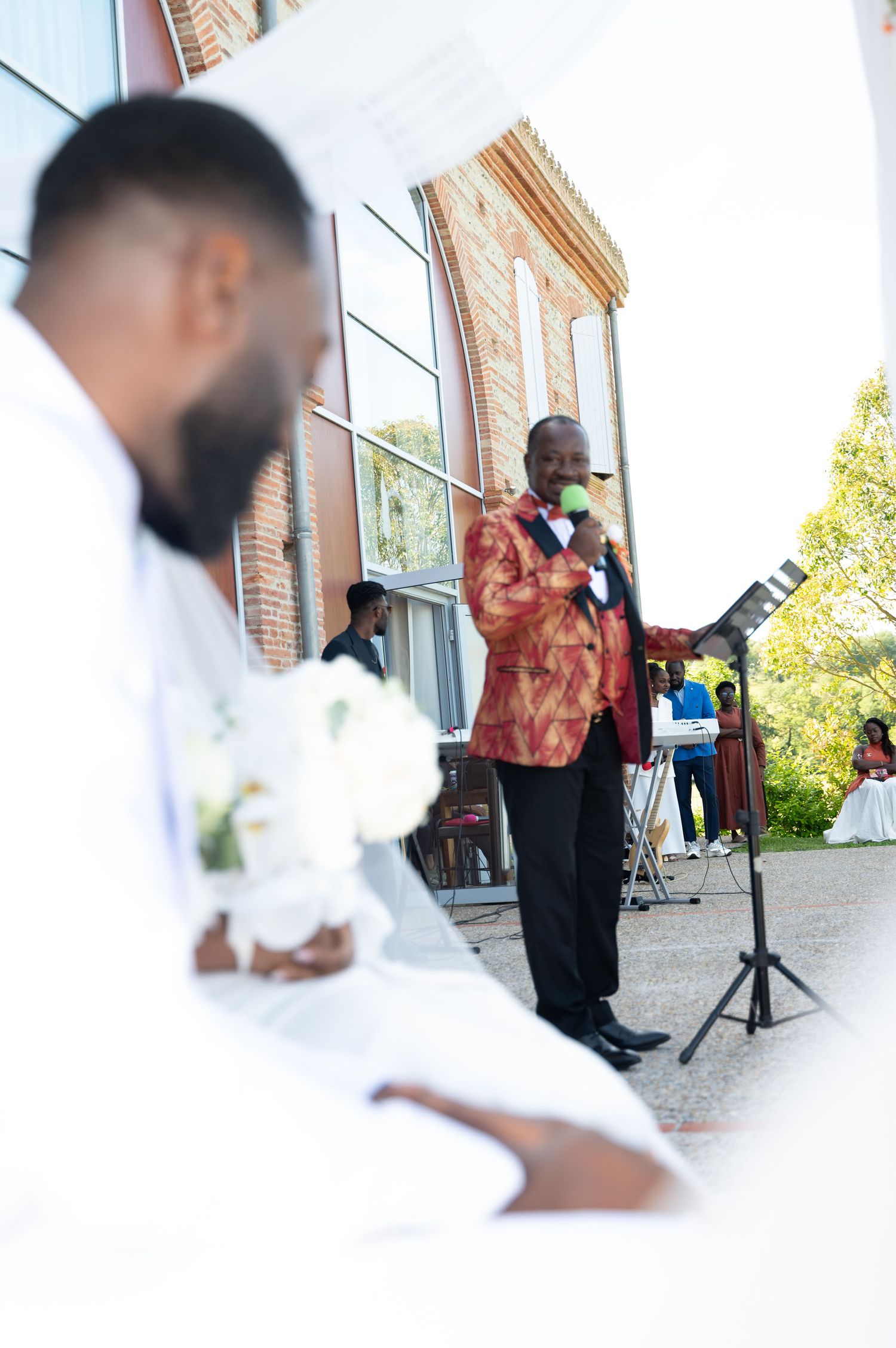 Man in red jacket sings at outdoor wedding ceremony with microphone and stand.