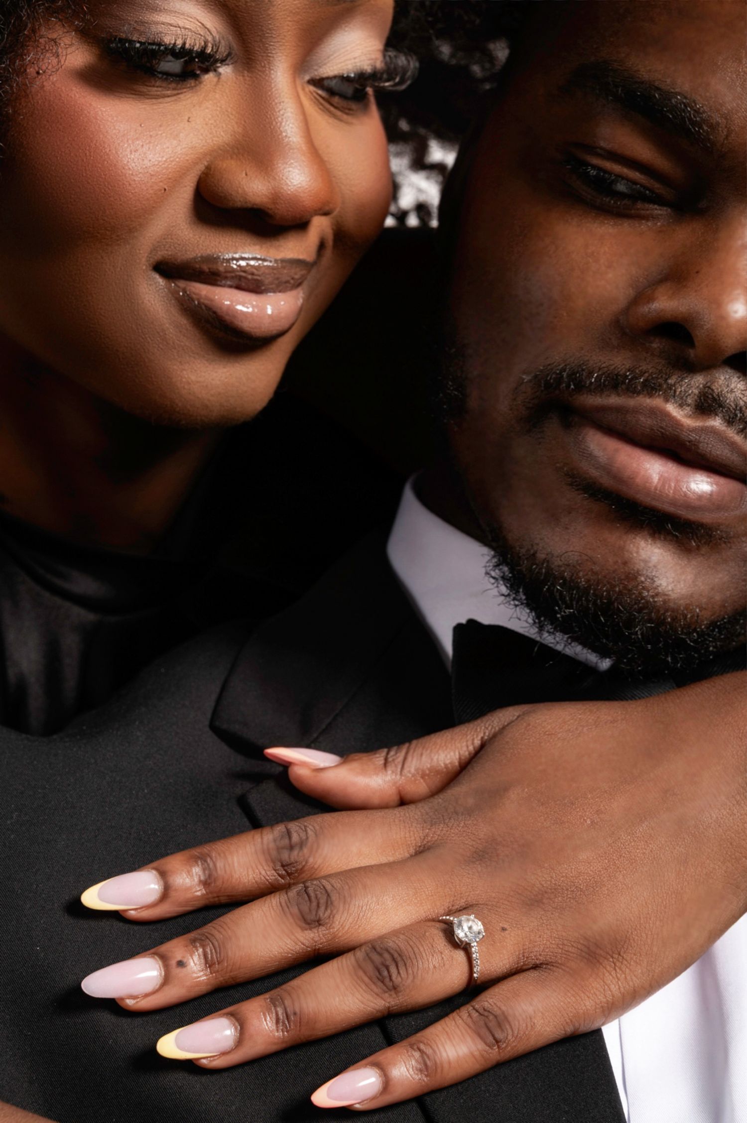 A couple in formal wear showcases an engagement ring with a diamond solitaire, her nails painted in yellow French tips.