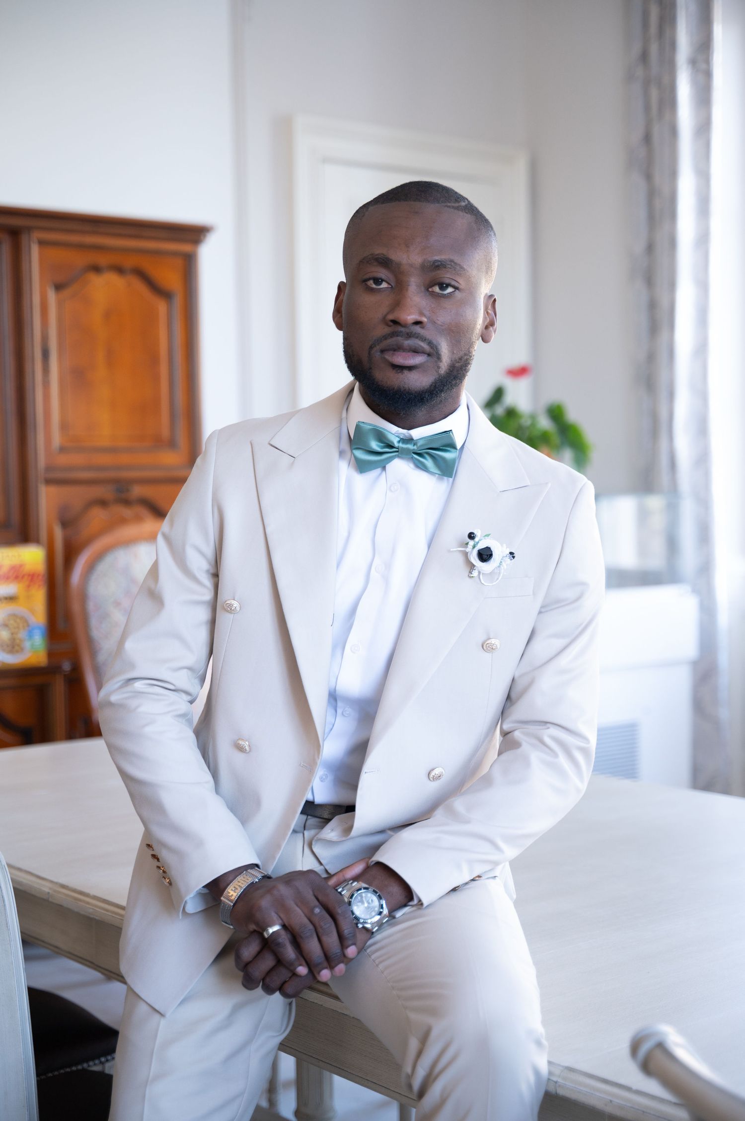 A well-dressed Black man in a cream double-breasted suit with a mint bow tie sits confidently at a table.