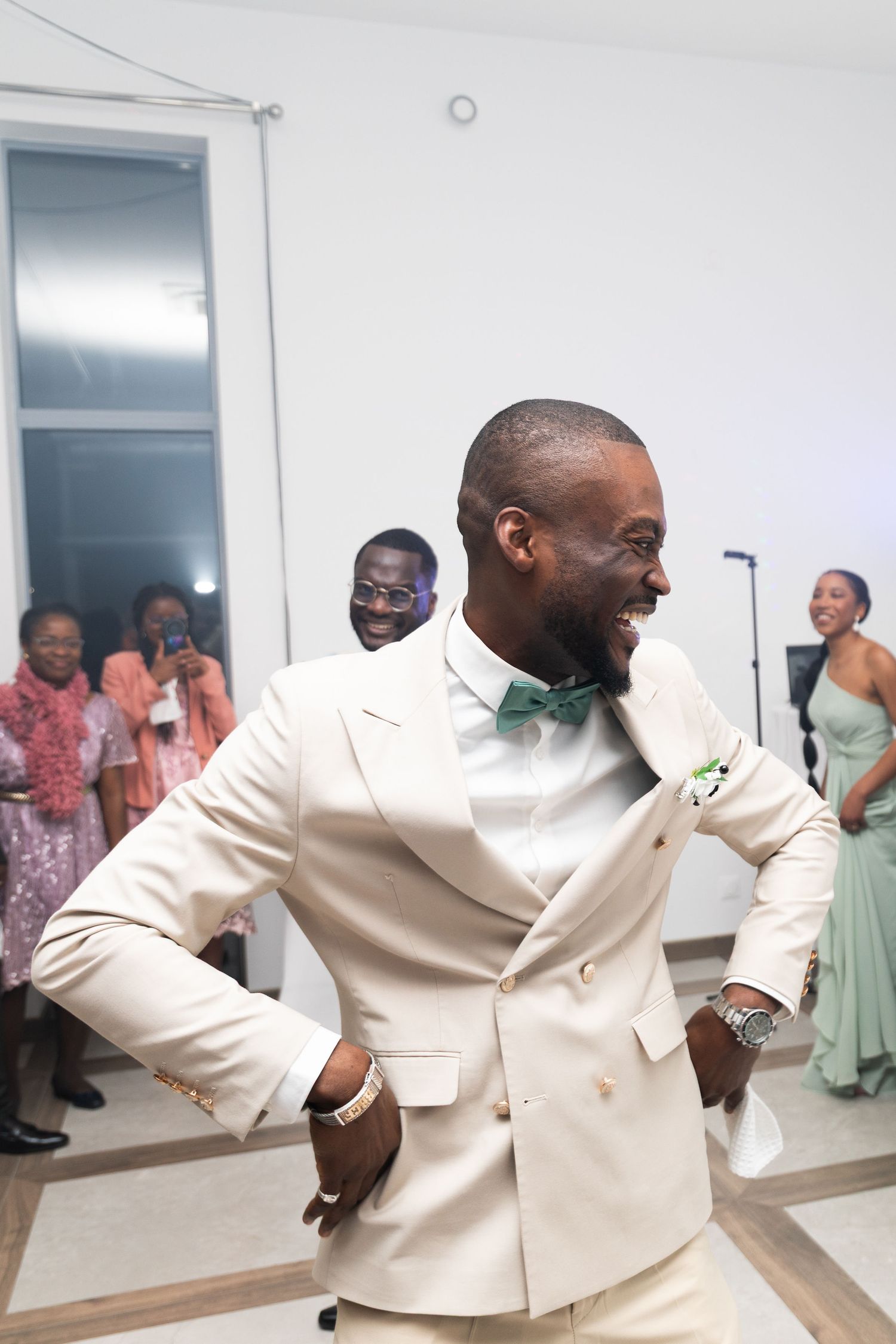 Joyful groom in cream suit with green bow tie dancing at wedding reception with guests watching.