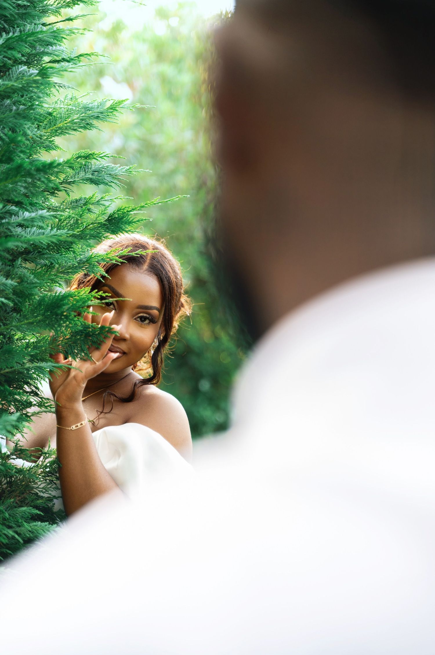 A bride in a white dress smiles shyly behind green foliage while her groom stands in the foreground.