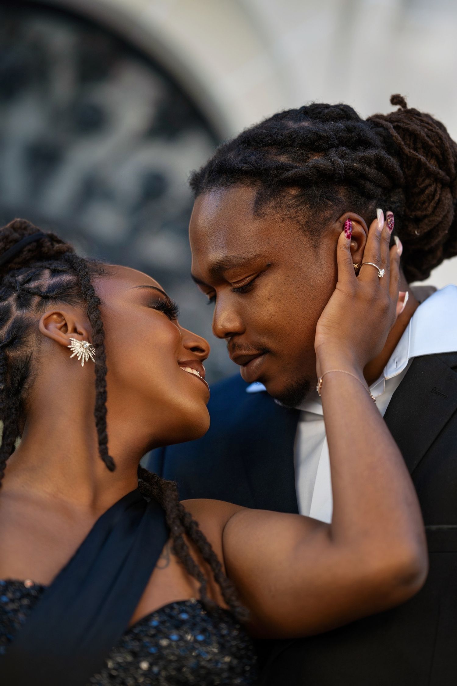 A couple shares a romantic moment, nearly kissing, both wearing elegant formal attire with dreadlocks hairstyles.