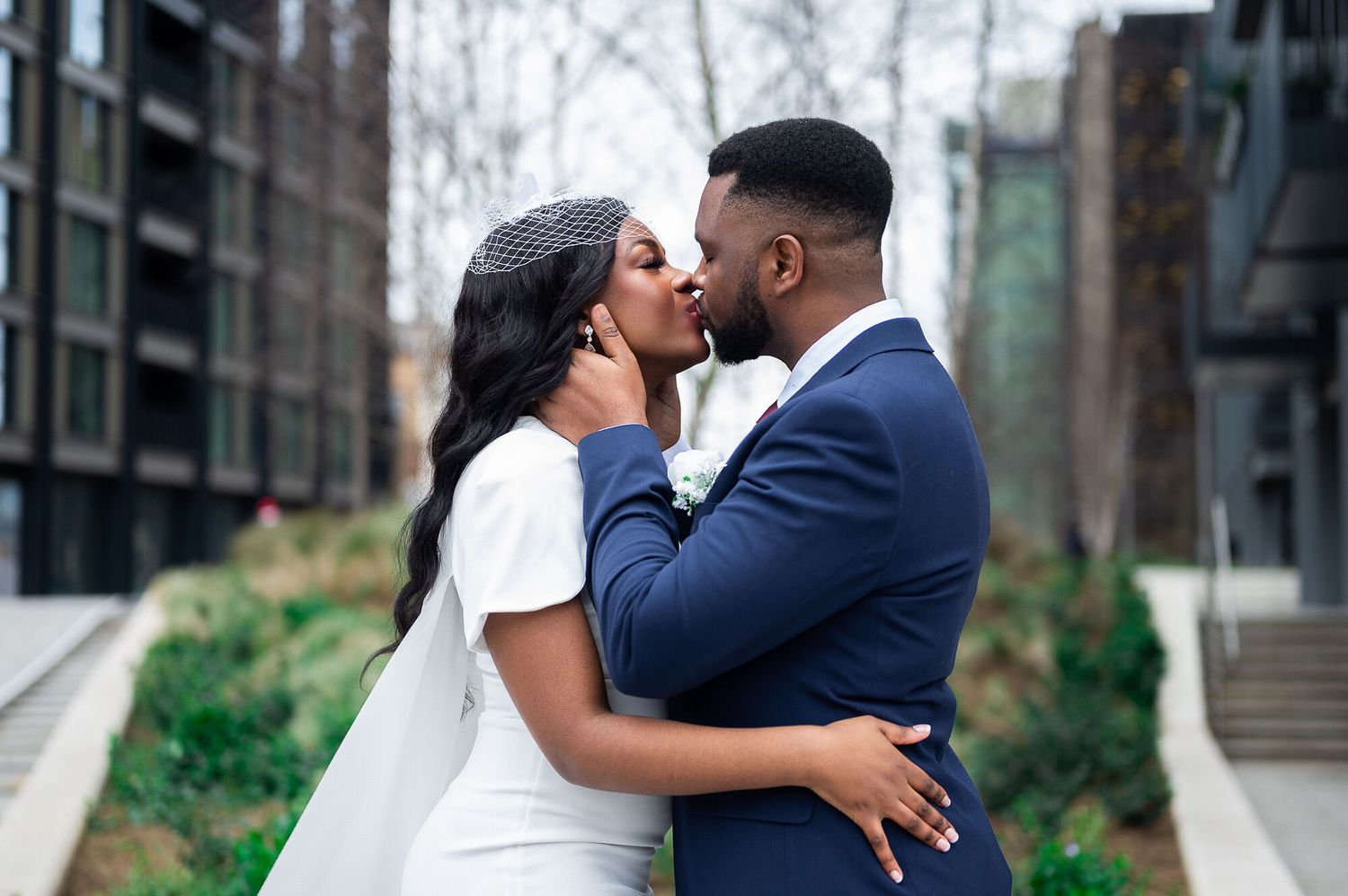 A Black couple shares a romantic kiss outdoors on their wedding day, groom in navy suit, bride in white.