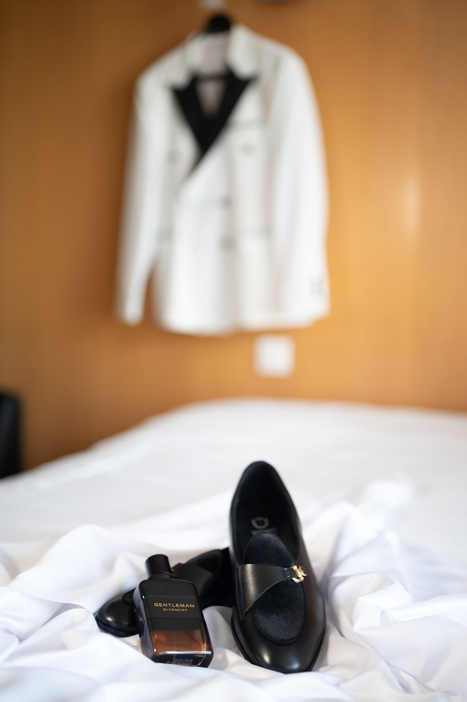 Black loafers and Givenchy Gentleman cologne on bed with white tuxedo jacket hanging in background.