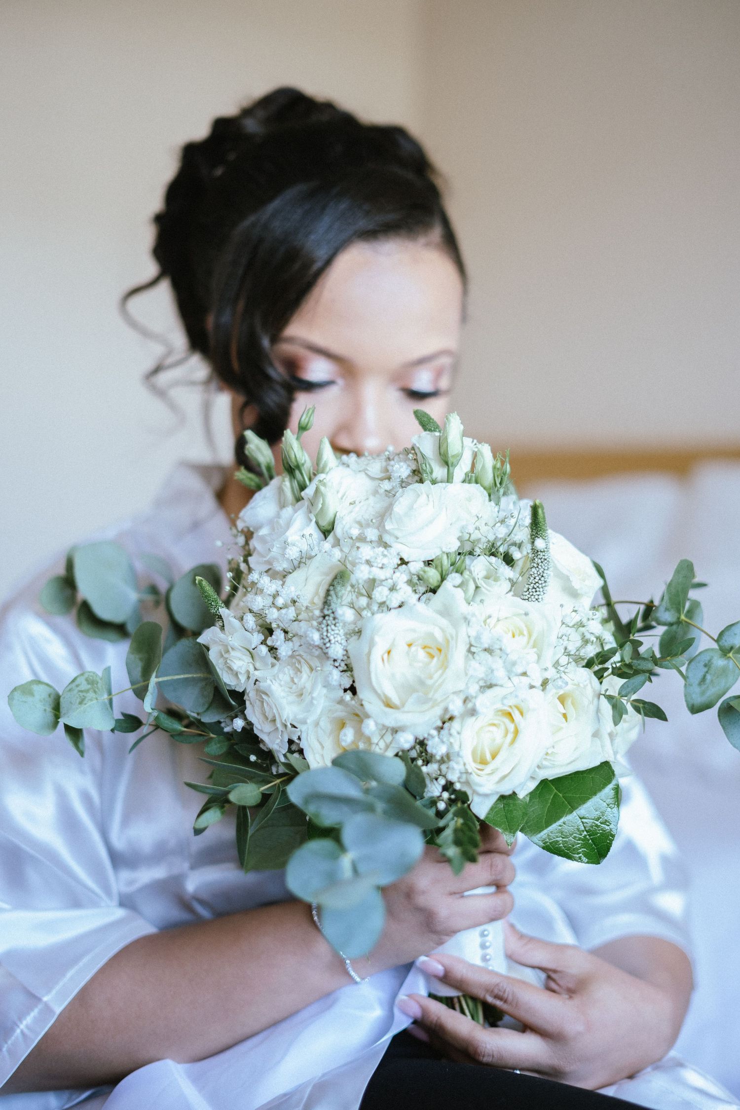 Bride in white satin robe holding a lush bouquet of white roses and eucalyptus to her face.