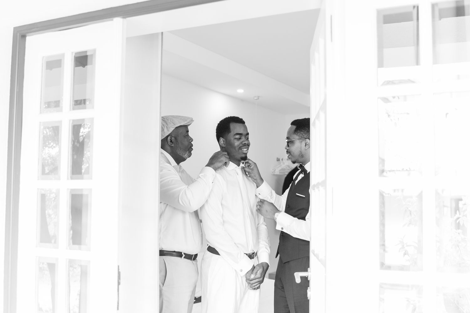 Black and white photo of three men helping groom get dressed in white suit in doorway before wedding.